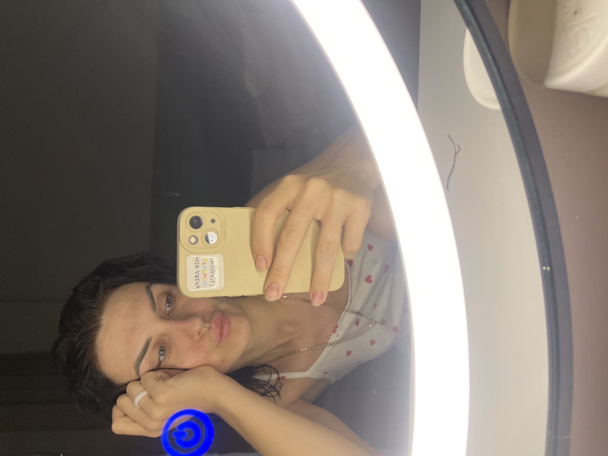 The image is a selfie taken in a mirror, showing a young woman with dark hair and light eyes. She is resting her head on her hand, with a slightly melancholic expression. She is wearing a white tank top with a small red heart pattern and a silver necklace. A yellow phone case is visible, held in her hand, with stickers on the back. One sticker has text that appears to be in Cyrillic script. The background is dark, with a prominent bright, circular light fixture, possibly a ring light, illuminating her face. A blue light ring is also visible in the bottom left corner of the image. The overall setting appears to be indoors, likely a bathroom or dressing area. The lighting suggests it might be evening or nighttime.