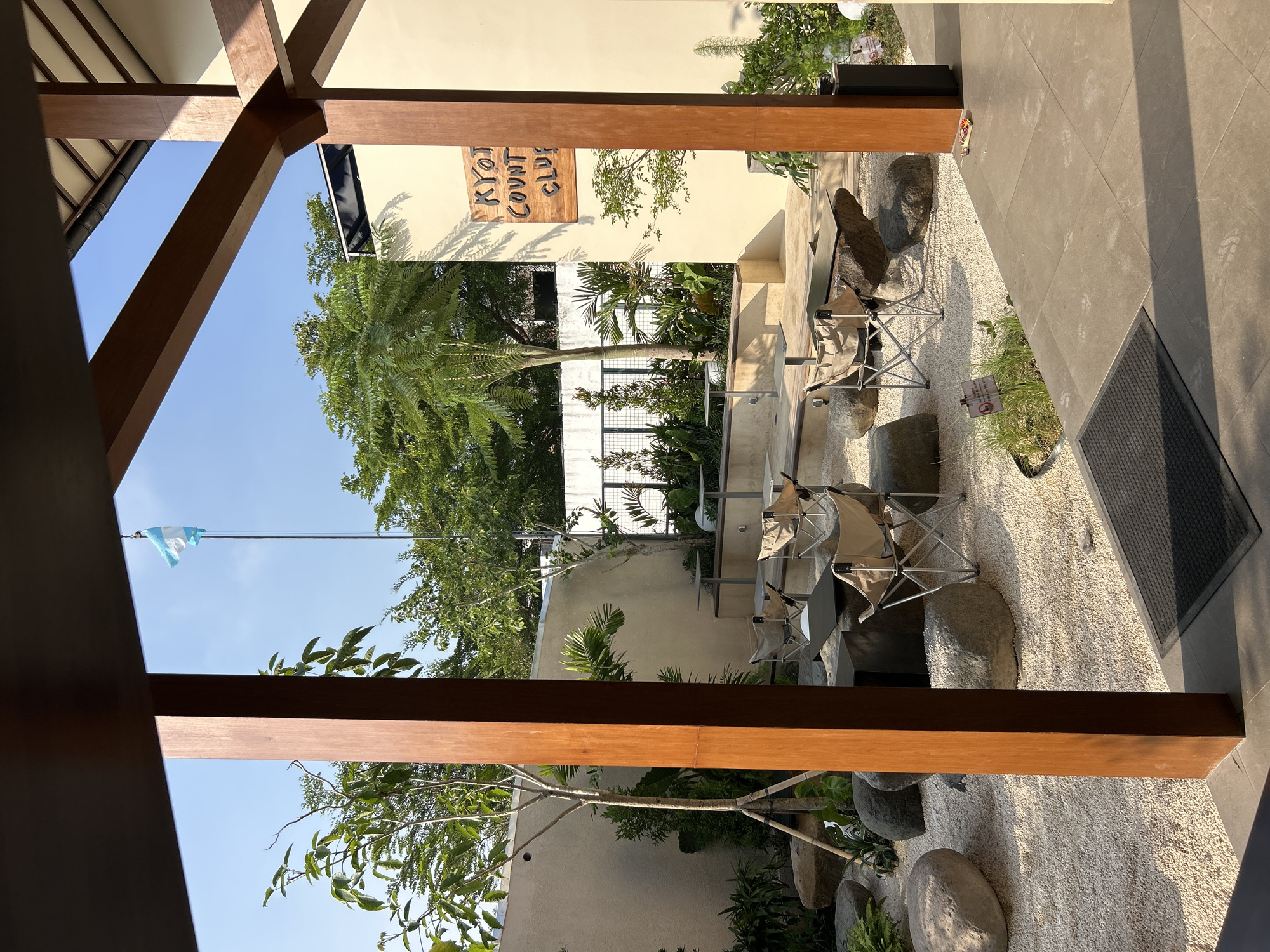 The image depicts an outdoor seating area at "Kyoto Country Club" in North Kuta, Indonesia. The space is designed with a Japanese garden aesthetic, featuring a light-colored gravel ground, large rounded stones, and various plants. Several portable camping chairs and small tables are arranged for patrons. In the background, a building with wooden architectural elements and a sign with the establishment's name is visible. A flag, appearing to be the flag of Argentina, flies from a flagpole. The scene is illuminated by natural sunlight, casting shadows and highlighting the textures of the environment. No people are present in the image.