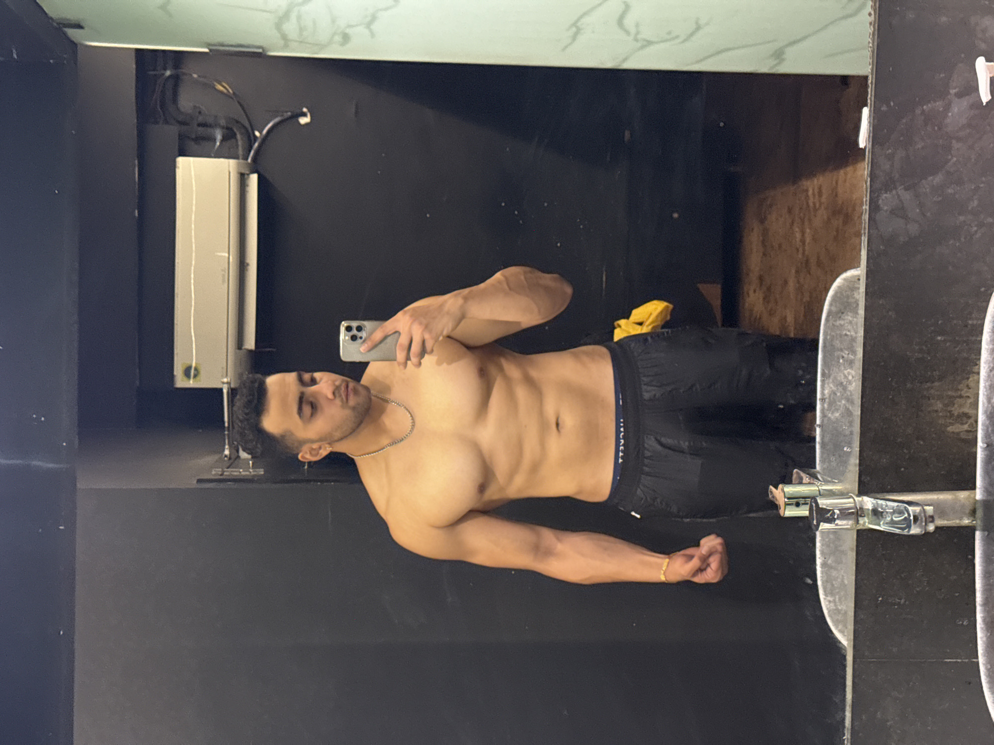 A man is taking a selfie in a bathroom mirror. The man has short, dark hair and a light skin tone. He is shirtless, wearing black shorts, a gold bracelet, and a silver necklace. The bathroom features dark gray walls, a light-colored countertop, and a mirror reflecting a black interior with a white electrical box. A yellow object is visible in the reflection. The location is Kolkata, India.