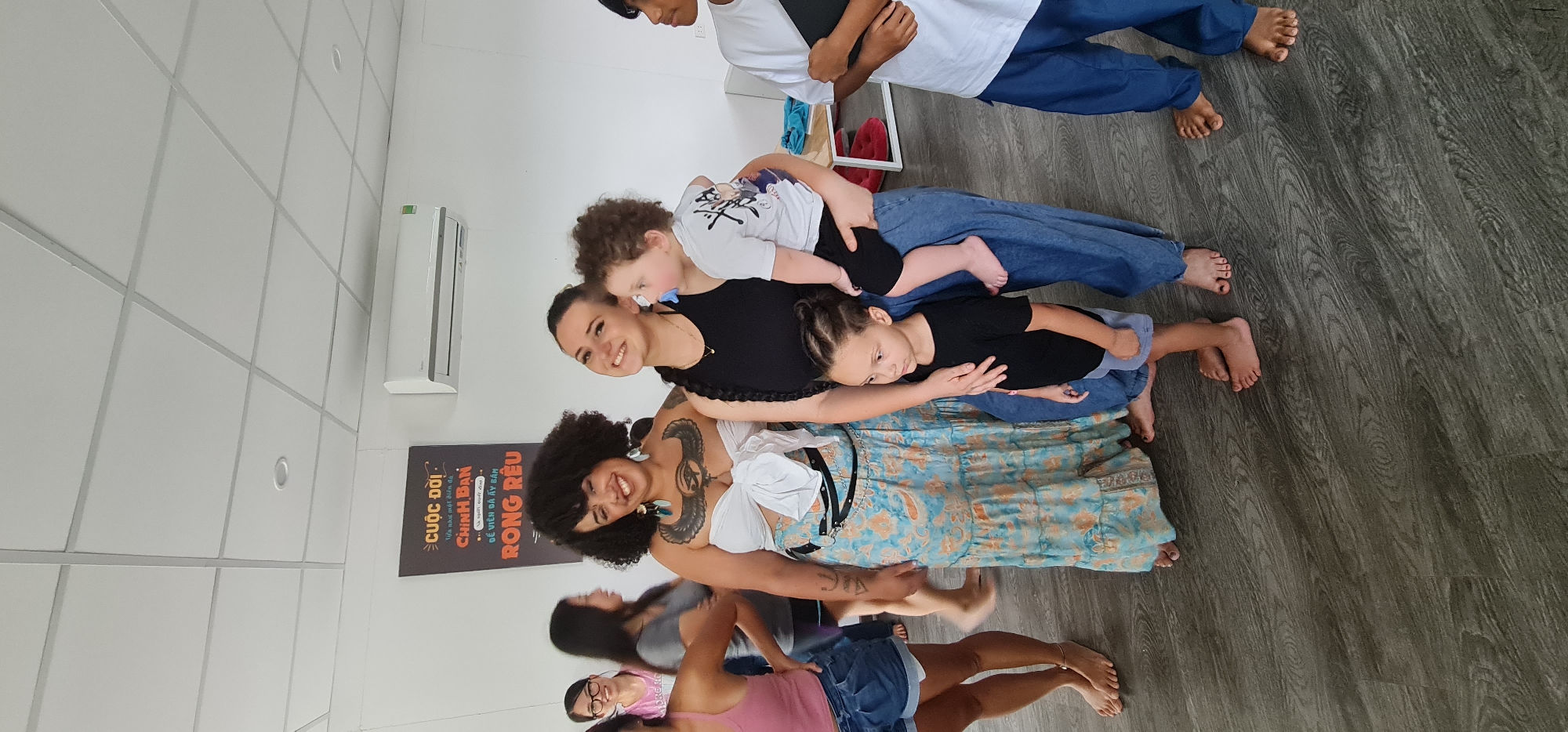 **Description:**
**Overview:** The image depicts a vertically oriented scene indoors, featuring a group of individuals aligned in a "human tower" arrangement. The setting appears to be a room with a modern design.
**People:**
- The group includes adults and children.
- A woman with dark curly hair and tattoos is at the base of the tower. Other women and children are stacked above her.
- The people are barefoot.
**Environment:**
- The room has a light-colored tiled ceiling and gray wood-look flooring.
- There is an air conditioning unit mounted on the wall.
- A sign is visible on the wall in Vietnamese. The sign includes the words "CUỘC ĐỜI," "CHÍNH BẠN," and "RONG RÊU."
**Objects:**
- The flooring has a wood-look pattern.
- A white object is visible near the top right of the frame.
**General:**
- The image shows a mix of skin tones.
- The orientation of the image suggests it may have been taken with a mobile device or similar.
- The sign visible may indicate that the location could be in Vietnam, potentially in the city of Đà Nẵng (based on the Vietnamese language).