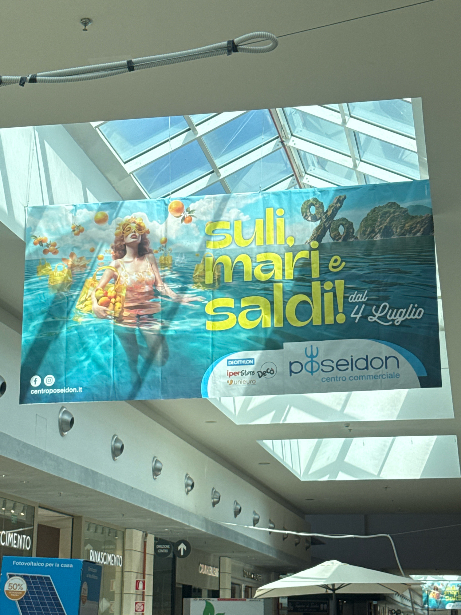This is a medium-distance, eye-level, horizontal shot of a large banner advertising a sale at the Poseidon Shopping Center. The banner features a woman in a swimsuit standing in water, surrounded by oranges and shopping bags. Text on the banner reads "SULI, mari e saldi! dal 4 Luglio". Below the banner, the Poseidon Shopping Center logo and Decathlon, IperStore Deco, and Unieuro logos are visible. The banner hangs from the ceiling of what appears to be an indoor shopping mall, with a skylight above and storefronts visible below. A solar panel advertisement is also visible in the lower left corner. The location is identified as Carini, Italy.