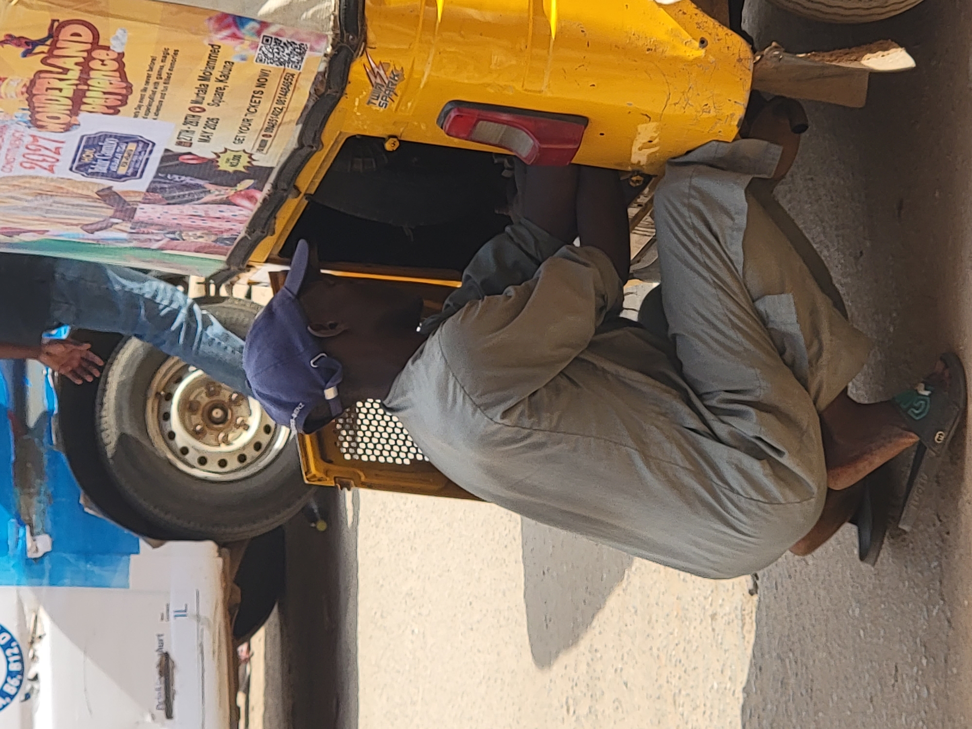 An outdoor, bright daylight scene in Maiburiji, Nigeria, depicts a man engaged in maintenance work on a yellow three-wheeled vehicle, resembling a tuk-tuk or auto-rickshaw. The man, dressed in a light-colored, possibly grey, long-sleeved shirt and pants, a blue cap, and black flip-flops, is bent over, examining the rear undercarriage of the vehicle with focused attention. A red taillight is visible on the yellow vehicle, which also bears a "TWIN SPARK" sticker. Part of another person, wearing blue jeans, is visible on the left side of the frame.
A large, somewhat faded advertisement poster is affixed to the side of the yellow vehicle. The poster promotes an event called "2027 WONDERLAND MAGIC!", scheduled for "27TH - 28TH MAY 2026" at "Murtala Mohammed Square, Kaduna." It encourages viewers to "GET YOUR TICKETS NOW!" and lists Nigerian phone numbers: "08084251422, 08144845550." Further details on the poster include descriptions like "Day event like never before! experienced with games, magic adventures and fun filled memories." A QR code and an image of a character resembling Spider-Man are also present.
The ground appears to be paved, possibly asphalt or concrete, with shadows suggesting clear, sunny weather. A car wheel is visible behind the working man. On the far left, partial views of white and blue containers are present, one displaying the text "86.812. D" and "Aurt." The man's blue cap has some faint white text, possibly "BENZ," and his flip-flops feature a green "G" logo.