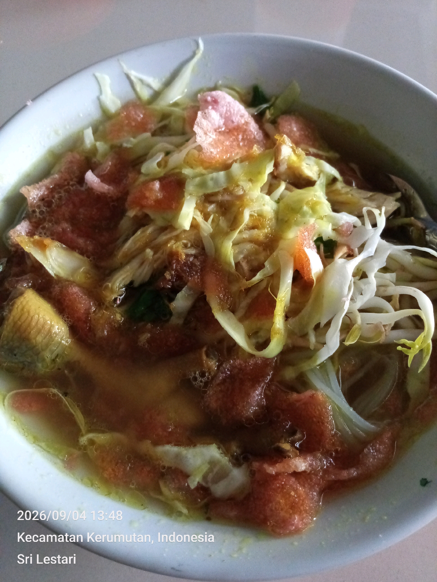 A close-up, overhead shot shows a bowl of Indonesian soup, likely Soto Ayam, filled with chicken, bean sprouts, shredded cabbage, and red crackers. The soup is a light yellow broth with visible oil slicks. The bowl is white and sits on a light-colored surface. Text overlays at the bottom left of the image read "2026/09/04 13:48 Kecamatan Kerumutan, Indonesia Sri Lestari". The image appears to be taken indoors with natural lighting. There are no people visible in the image.