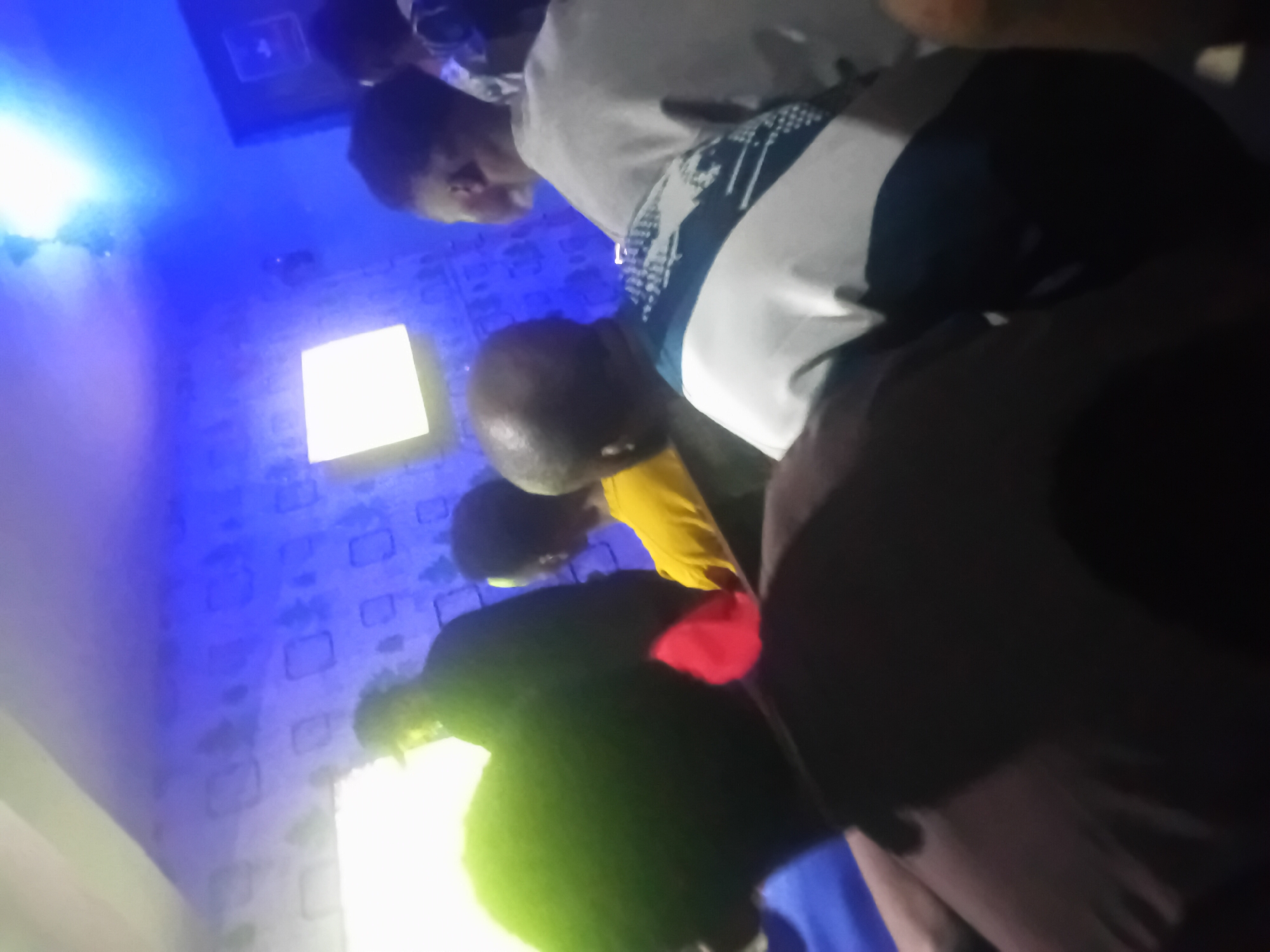 A group of people are gathered indoors in a room with blue patterned walls. One person is wearing a white shirt with black stripes and a bald head, while another is wearing a yellow shirt. Other people are wearing dark clothing. The scene is illuminated by artificial light sources, including a square light fixture. The location appears to be Lokoja, Nigeria, as specified in the request.