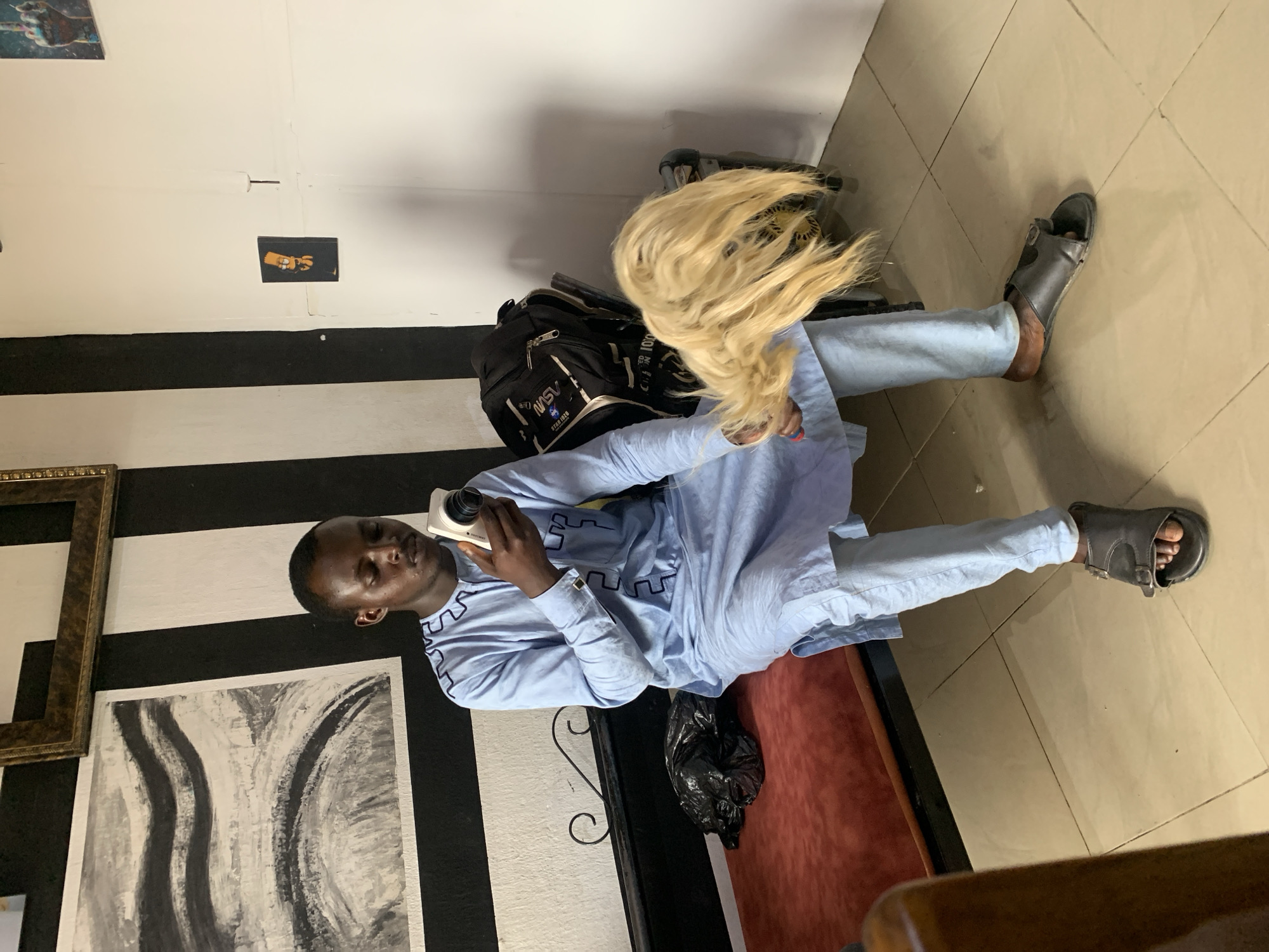 A man is seated on what appears to be a low couch or bench, holding a smartphone up to his face as if taking a picture or recording video. He's wearing a light blue, long-sleeved shirt and light blue pants with grey sandals. Adjacent to the man are objects including a large, unkempt, light-blonde wig and a black backpack. The background includes a wall with alternating black and white stripes, as well as a framed picture and an abstract art piece. Another small picture hangs near the ceiling. A black trash bag can be seen near the couch, and the floor is tiled. The location appears to be indoors. I cannot ascertain the specific location within Jos, Nigeria, or other details beyond what is visible in the image.
