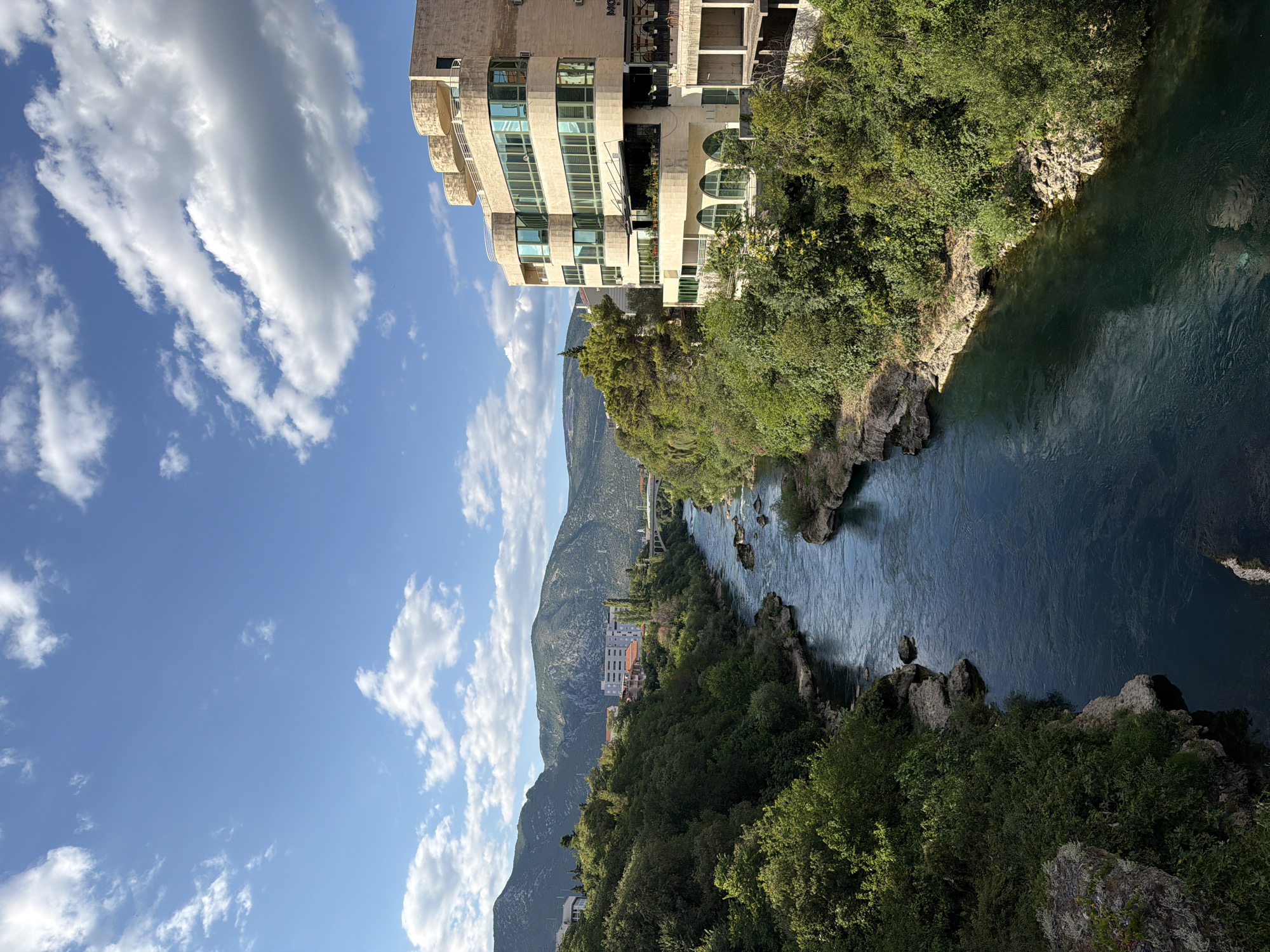 This image, taken from a high vantage point in Mostar, Bosnia and Herzegovina, captures a scenic view of the Neretva River flowing through a verdant gorge. The river, a deep, tranquil blue, meanders through rocky outcrops and is flanked by lush green trees that cover the steep slopes. In the background, imposing mountains rise under a bright blue sky dotted with fluffy white clouds.
On the left side of the frame, a modern, multi-story building with a cream-colored facade stands prominently. Its architecture features curved balconies and large windows, contrasting with the natural landscape. The building appears to be situated on the edge of the gorge. Further into the distance, along the riverbank, glimpses of other buildings and infrastructure, possibly a bridge, can be seen, hinting at the presence of the city.
The orientation of the image suggests it was taken looking down and to the side. The lighting indicates it is daytime, likely mid-morning or afternoon, with clear weather. The overall impression is one of a serene and picturesque natural setting integrated with urban development. No people are visible in the image. There is some faint text on the building, including what appears to be "MO" on a sign.