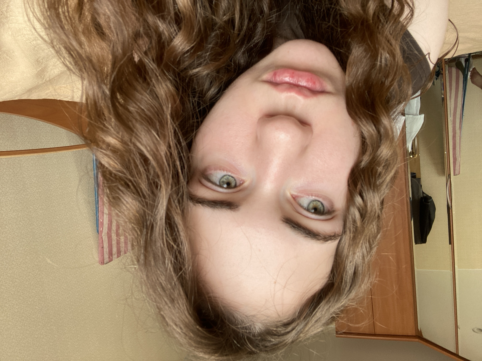 The image shows a close-up of a young woman's face, angled slightly to her left. She has long, wavy brown hair that frames her face. Her eyes are a striking green, with one slightly more open than the other, and she has defined eyebrows. Her lips are slightly parted, with a pouty expression. Her skin appears fair and smooth.
In the background, to the left, is a wooden wardrobe with mirrored doors, reflecting a closet area with clothing. To the right, a textured beige wall is visible, with a hint of a striped object, possibly a towel or curtain, hanging on it. A wooden headboard or edge of a bed is also visible on the far right.
The scene suggests an indoor setting, likely a bedroom or dressing area. There is no indication of the city of Zavodoukovsk, Russia, or any other specific geographical location. The image is a portrait of the woman, with the background providing context for the environment.