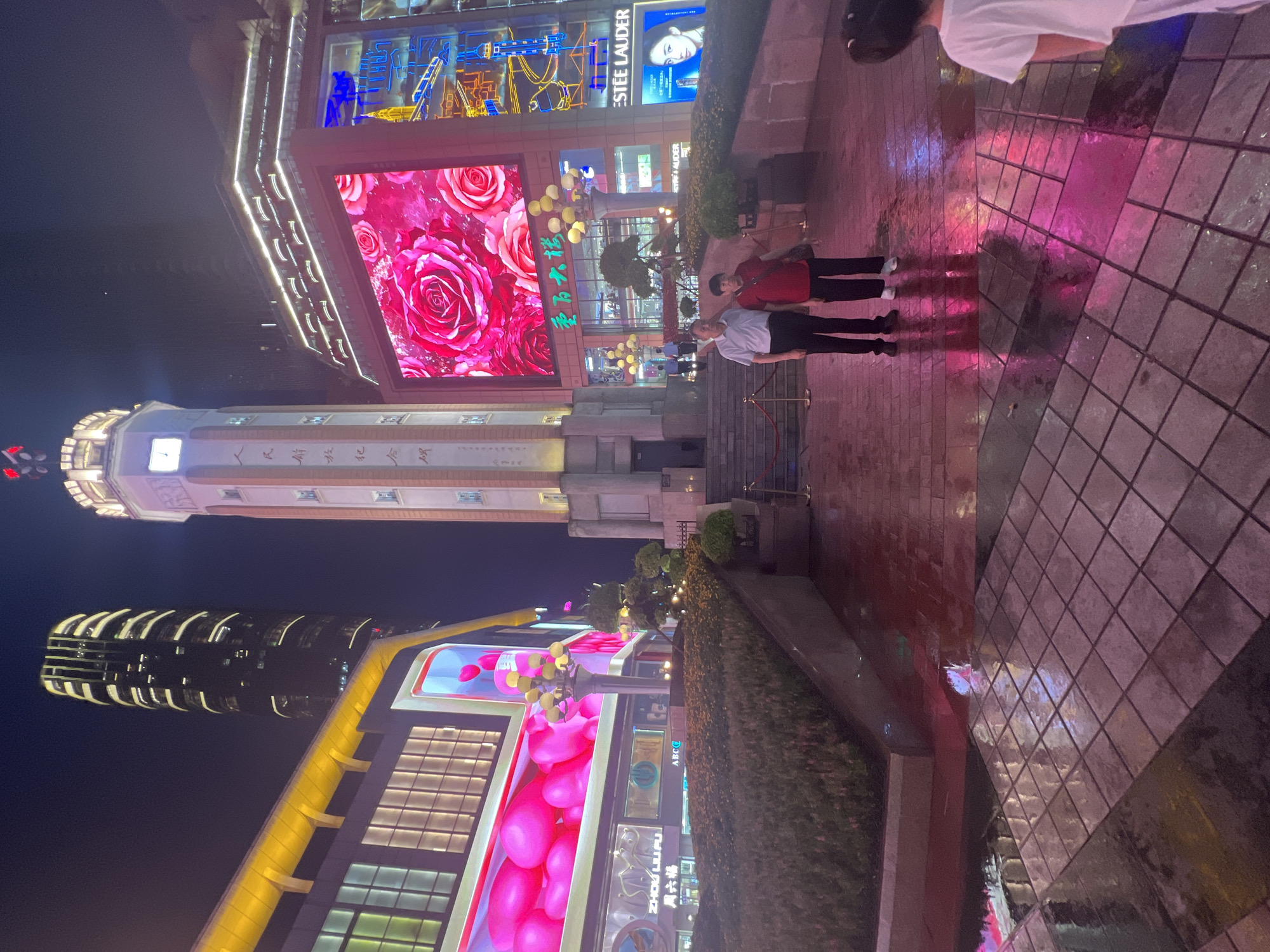 This image captures a night scene in Yuzhong District, China. A tall, modern skyscraper with illuminated vertical stripes stands on the left. To its right is a clock tower, featuring traditional Chinese writing on its sides. In the foreground, a couple stands on a wet plaza, their reflections shimmering on the ground from the vibrant pink and purple lights of the surrounding buildings. The plaza is paved with square tiles, and a landscaped garden with flowering bushes is visible. A large LED screen displays a close-up image of pink roses. The buildings are adorned with bright, colorful lights, creating a festive and urban atmosphere. The scene is illuminated by artificial lights, casting reflections on the wet ground, suggesting it has recently rained.