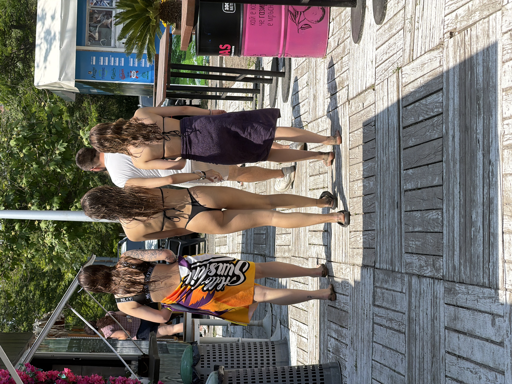 The image depicts an outdoor scene on a sunny day, likely a boardwalk or beach area, identified as being in Burgas, Bulgaria, based on the Cyrillic text visible on signs and barrels.
In the foreground, a group of four individuals, consisting of three women and one man, walk away from the camera. All three women have long, wet, dark hair.
1. The leftmost woman wears a black sports bra with a white band displaying partial text, and a multi-colored towel (orange, yellow, purple, white) wrapped around her waist, featuring the text "Hello Sunshine." A floral tattoo is visible on her left upper arm. She is barefoot, with sand on her feet.
2. The central woman wears a black string bikini top and a black thong bikini bottom. She is barefoot, with sand on her feet.
3. Behind and to the right of the central woman, a man in a white t-shirt and light-colored shorts is partially visible, wearing white sneakers.
4. The rightmost woman wears a black string bikini top and a dark purple towel wrapped around her waist. She is barefoot, with sand on her feet.
To the far left, a food or beverage stall with a dark green facade and an extended awning is adorned with pink artificial flowers. Inside, a woman in a plaid shirt and dark shorts is visible. Two dark gray and green perforated trash bins stand nearby.
To the far right, a blue kiosk with a white awning advertises various drink brands including "Zatecky," "TUBORG," and "Carlsberg," with prices listed in Bulgarian Cyrillic. A small potted palm plant is positioned in front of the kiosk. Dark brown wooden tables with black metal bases are visible. Further right, two stacked pink barrels, functioning as bins, display Cyrillic text and a fruit illustration. A gray utility pole stands between the kiosk and the barrels.
The ground is a light-colored wooden deck constructed from square panels with alternating plank directions, featuring shadows from the people and structures. Small weeds grow in some gaps between the planks. The background consists of lush green trees and foliage under a clear blue sky. A white car is partially visible in the far background.