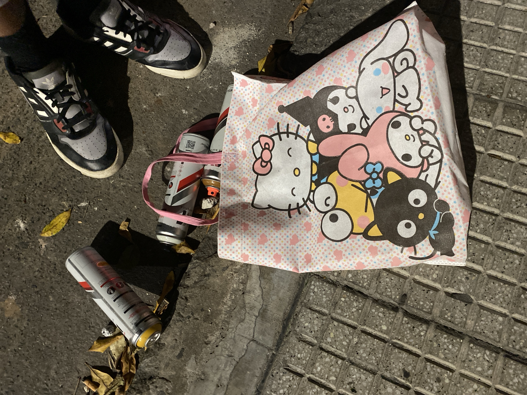 A street scene at night. The ground is a mix of asphalt and concrete sidewalk, with a section of tiled pavement with square patterns. There are two pairs of shoes, one on a person whose black-socked foot is visible, and another pair nearby. Next to the shoes is a tote bag with a light-pink heart pattern, featuring cartoon characters including Hello Kitty. Several used aerosol cans, possibly spray paint, are scattered around the bag and under the shoes. The cans are white with red and black markings. Some leaves are also scattered around. The exact location within Buenos Aires, Argentina, is not discernible based on the image alone.