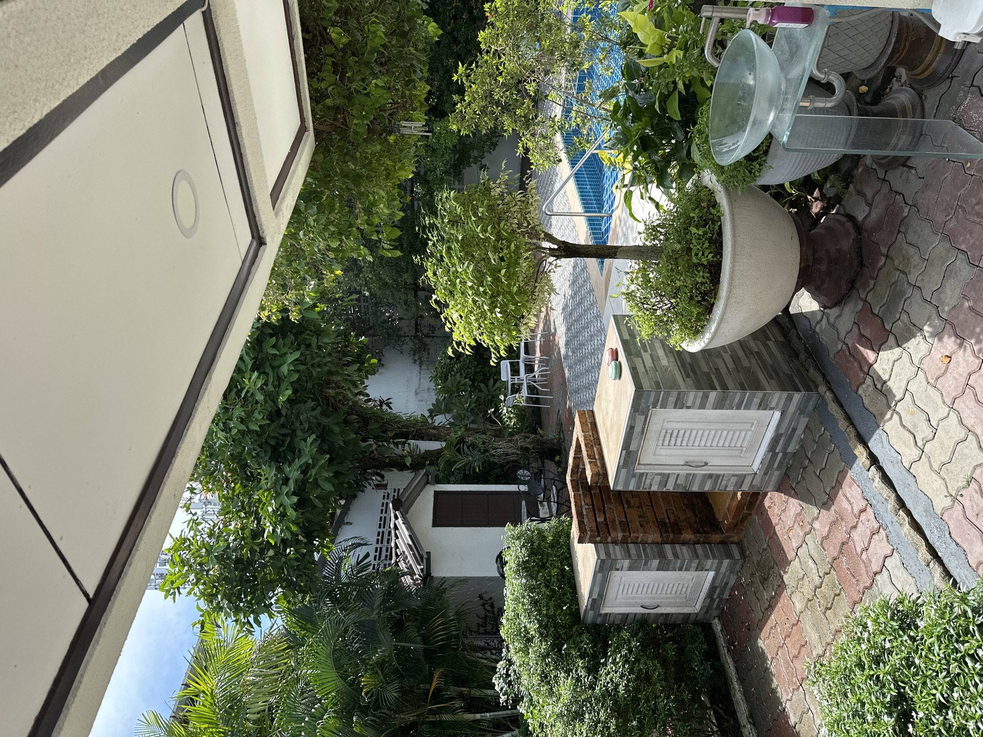The image shows a backyard patio area with a swimming pool. In the foreground, there are two outdoor cabinets with white doors, built next to a brick structure. Adjacent to these cabinets is a large potted plant and a glass sink with a faucet and soap dispenser. Beyond this, a swimming pool with blue tiles is visible, surrounded by lush green plants. In the background, there is a small white shed with a brown door and a white tiled roof. White outdoor chairs and tables are also visible near the shed and by the pool. The ground is paved with interlocking bricks of red, gray, and beige colors. The scene appears to be set in a tropical or sub-tropical climate, with dense foliage and palm trees present. The overall impression is that of a well-maintained outdoor living space.