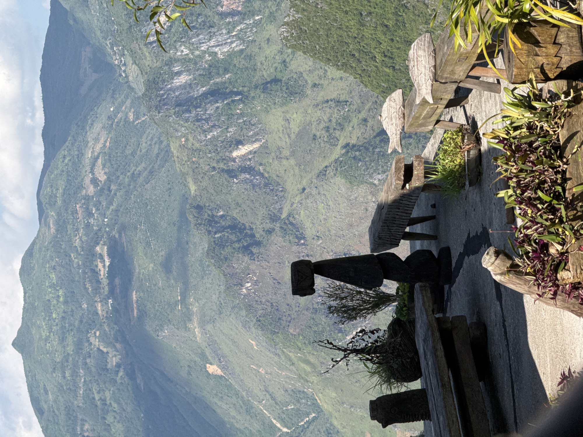 The image shows an outdoor scene with a mountainous landscape as the backdrop. A seating area is constructed on the edge of a platform, likely a viewpoint. The seating area includes wooden benches and planters with greenery. The mountains are covered in vegetation, creating a lush, green environment. The location is likely Đồng Văn Commune, Vietnam, suggested by the typical landscape of the region. A stone sculpture is visible in the foreground.