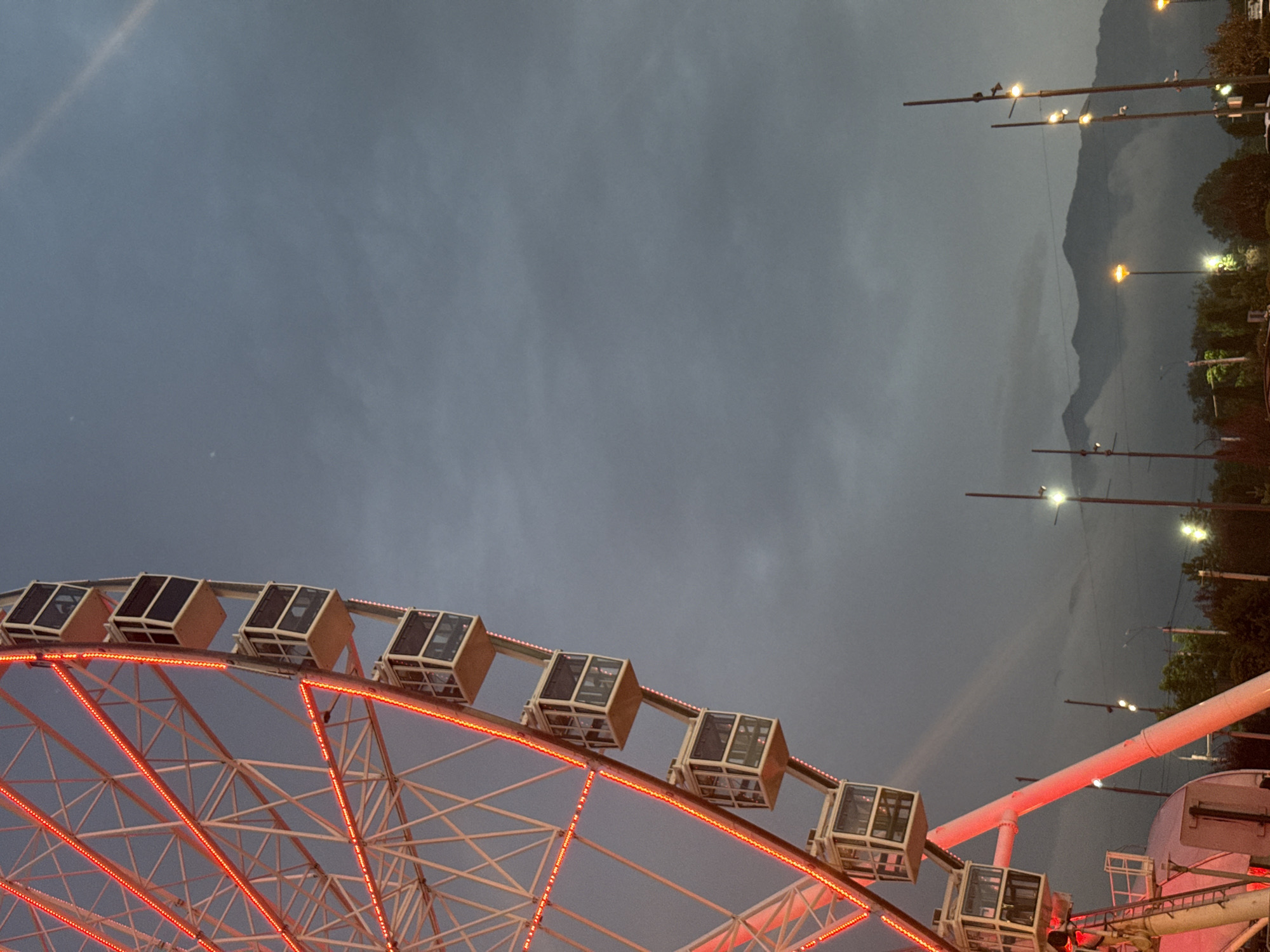 The image shows a Ferris wheel lit with red lights at dusk. Several gondolas are visible on the wheel, which is white with red accents. In the background, mountains are shrouded in clouds, and several streetlights are on. The sky is a dark, cloudy gray. This scene is characteristic of the area around the Kok Tobe hill in Almaty, Kazakhstan, a popular spot with a Ferris wheel and panoramic views of the city and surrounding mountains. There are no people or specific actions depicted.