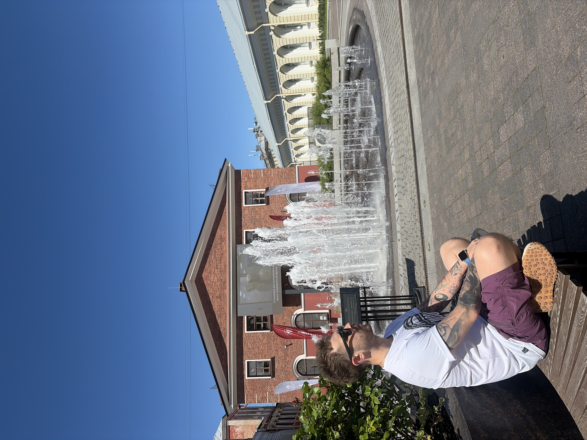 The image captures a sunny urban landscape in Saint Petersburg, Russia, featuring the Fountain "Peter I." In the foreground, a Caucasian male, appearing to be in his 20s or 30s, is seated cross-legged on a wooden slatted bench, positioned to the left of the frame. He has short brown hair, wears black sunglasses, a white short-sleeved t-shirt with a dark graphic design, reddish-purple shorts, and light-colored sneakers with textured soles. His arms are extensively tattooed, and he wears a blue-strapped watch on his left wrist and a thin gold chain. He is gazing towards the right, past the centrally located circular fountain, which is actively spraying multiple jets of water.
Behind the fountain, a two-story red brick building with a classical pediment and a dark roof is visible. A large outdoor screen is mounted on its facade, displaying an image and Russian text, partially identifying "Стажировка для школьников" (internship for schoolchildren). To the right of the red brick building, a long, light yellow building with a series of arched window recesses and a light-colored roof extends into the background. On the far left, partially obscured by green bushes, is a portion of another older brick building. The ground is paved with a pattern of square and rectangular grey and reddish-brown cobblestones. The sky is clear and deep blue, and bright sunlight casts pronounced shadows across the scene, indicating a clear day. Several promotional banners, predominantly red and white, are visible near the red brick building.