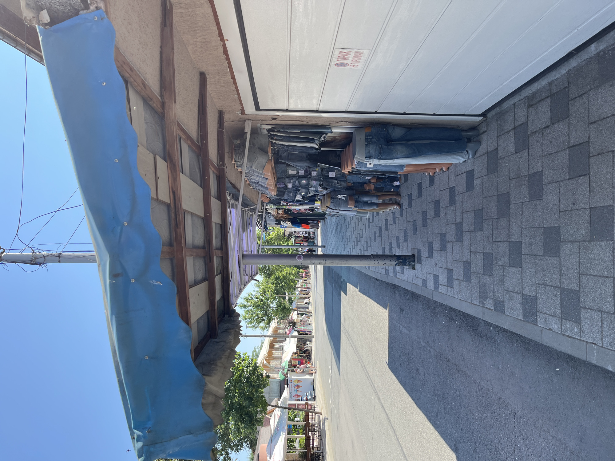 A street scene in Obzor, Bulgaria, shows a commercial area with shops and stalls lining a sidewalk next to a road. The foreground is dominated by a row of mannequins dressed in denim shorts and jeans displayed outside a shop. The shop has a large white garage door and a blue awning extending over the display. The sidewalk is paved with gray and dark gray square tiles. The road is asphalt and appears to be a street with some traffic. In the background, more shops and trees are visible, suggesting a busy pedestrian area. The sky is clear and blue, indicating a sunny day. A sign on the garage door is partially visible, with Cyrillic text.