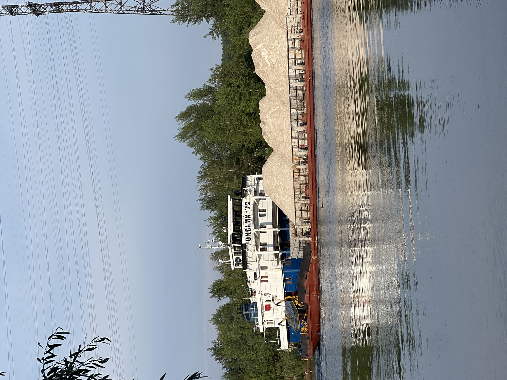 The image shows a barge filled with a large pile of light-colored material, possibly sand or gravel, being towed on a body of water. A tugboat is attached to the front of the barge, pushing it. The tugboat has the text "ОКСКИЙ-72" visible on its side. The scene is set against a backdrop of lush green trees lining the shore. In the distance, a tall power line tower and multiple electrical wires stretch across the sky. The sky is clear and pale blue, suggesting it is daytime, likely late afternoon or early evening due to the warm, golden light reflecting on the water and illuminating the side of the barge. The water's surface is calm with gentle ripples. The overall impression is one of industrial transport and a serene natural landscape. The orientation of the image is landscape with the barge and its cargo running from the bottom left to the top right of the frame.