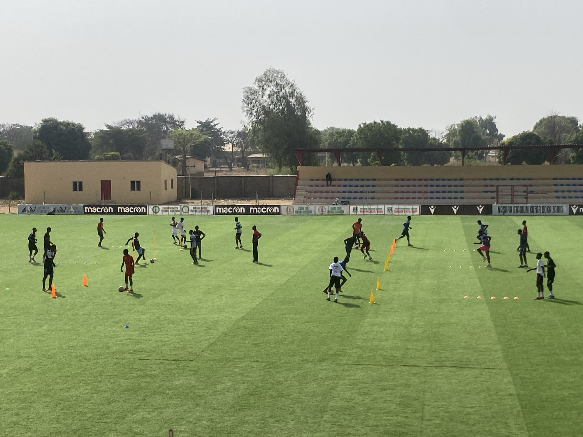 The image depicts a soccer field, likely located in Tudun Wada, Nigeria, at the Nasara Stadium Kofar Doka Zaria. A group of soccer players are seen practicing on the green field, utilizing orange and yellow cones as training aids. The backdrop includes a low building, a perimeter fence, some trees, and a small stadium seating area, part of which is visible. Logos of "macron" are displayed on a banner bordering the field. The sky is overcast, suggesting a hazy or dusty atmospheric condition.