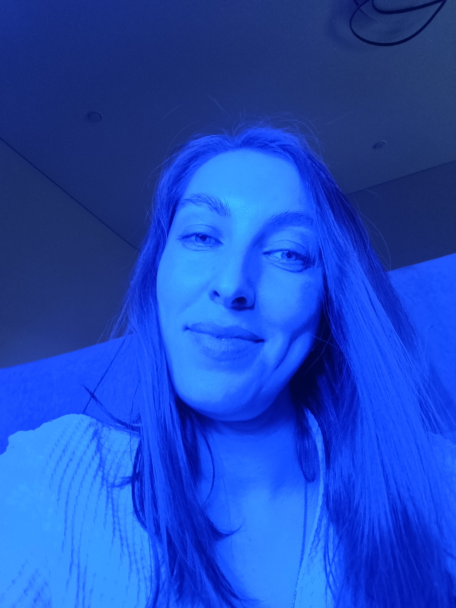 This image is a close-up portrait of a young woman, likely a selfie, taken indoors under strong blue lighting. The woman, who appears to be in Chaikovsky, Russia, has long, dark hair that is also illuminated by the blue light. She is looking slightly upwards and to the right, with a soft, subtle smile on her face, revealing a slight dimple on her right cheek. Her eyes are a light color. She is wearing a light-colored, possibly white or cream, textured top or sweater, and a delicate silver-colored necklace is visible around her neck.
The background is predominantly dark blue, consistent with the pervasive blue lighting, and appears to be a wall or ceiling. Hints of architectural details are visible, including what looks like a small circular fixture or vent in the upper left, and faint lines that could be seams or edges of panels. There are also some indistinct dark lines in the upper right, possibly wires or other fixtures. The intense blue illumination gives the scene a cool, possibly nocturnal or club-like atmosphere. No specific activity beyond posing for the photograph is depicted. There is no visible text in the image. The overall mood conveyed is calm and somewhat ethereal due to the distinct lighting.