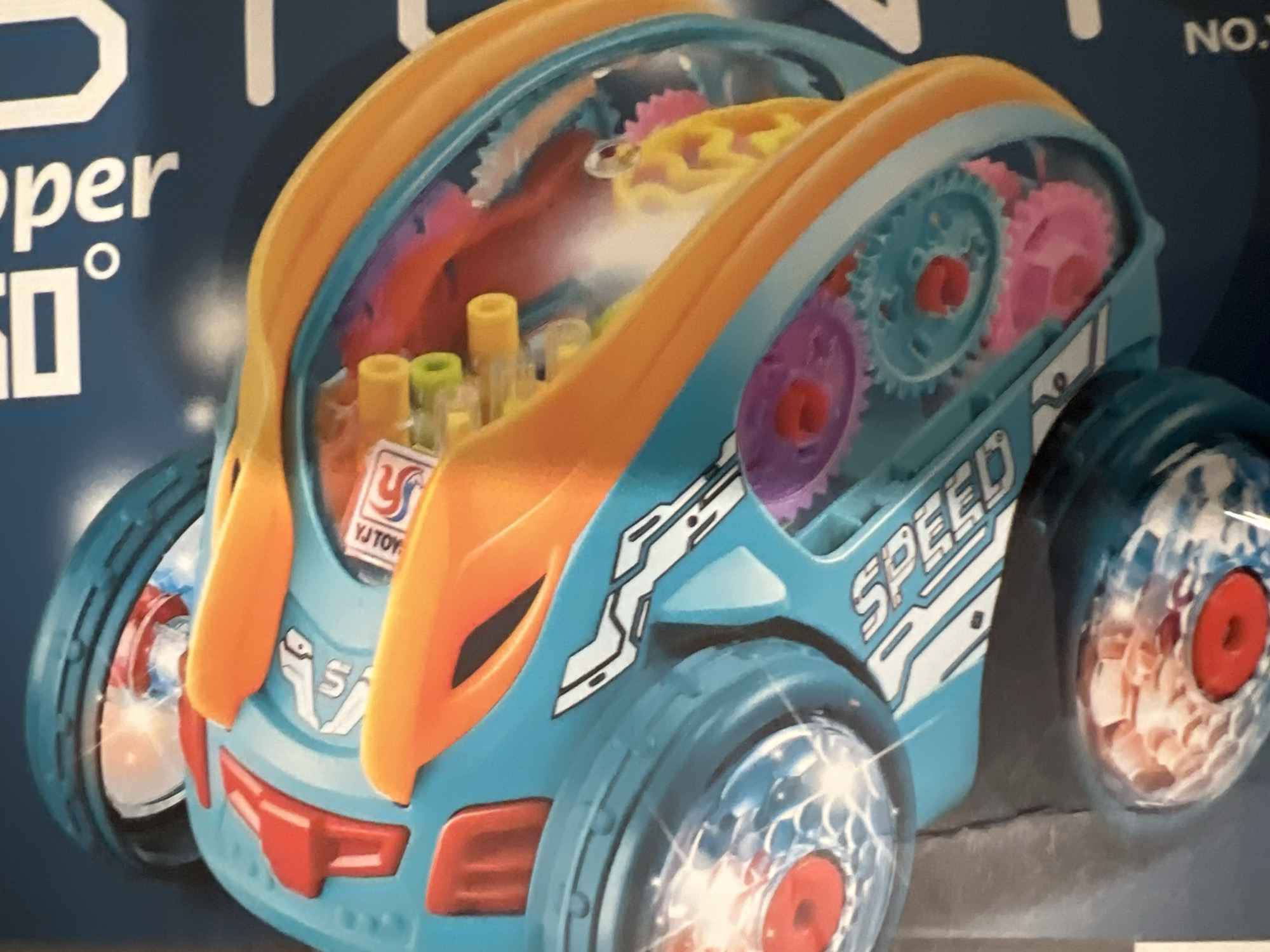 The image shows a close-up of a blue and orange toy with visible gears and colorful lights. The toy has "SPEED" written on its side in white lettering. Text on the blue packaging in the background includes the word "SUPER" in large white letters, and "NO." vertically on the right side of the frame. The toy is likely a vehicle or a mechanical device with transparent sections revealing internal workings. There is no indication of the city of Yekaterinburg, Russia, in the image.