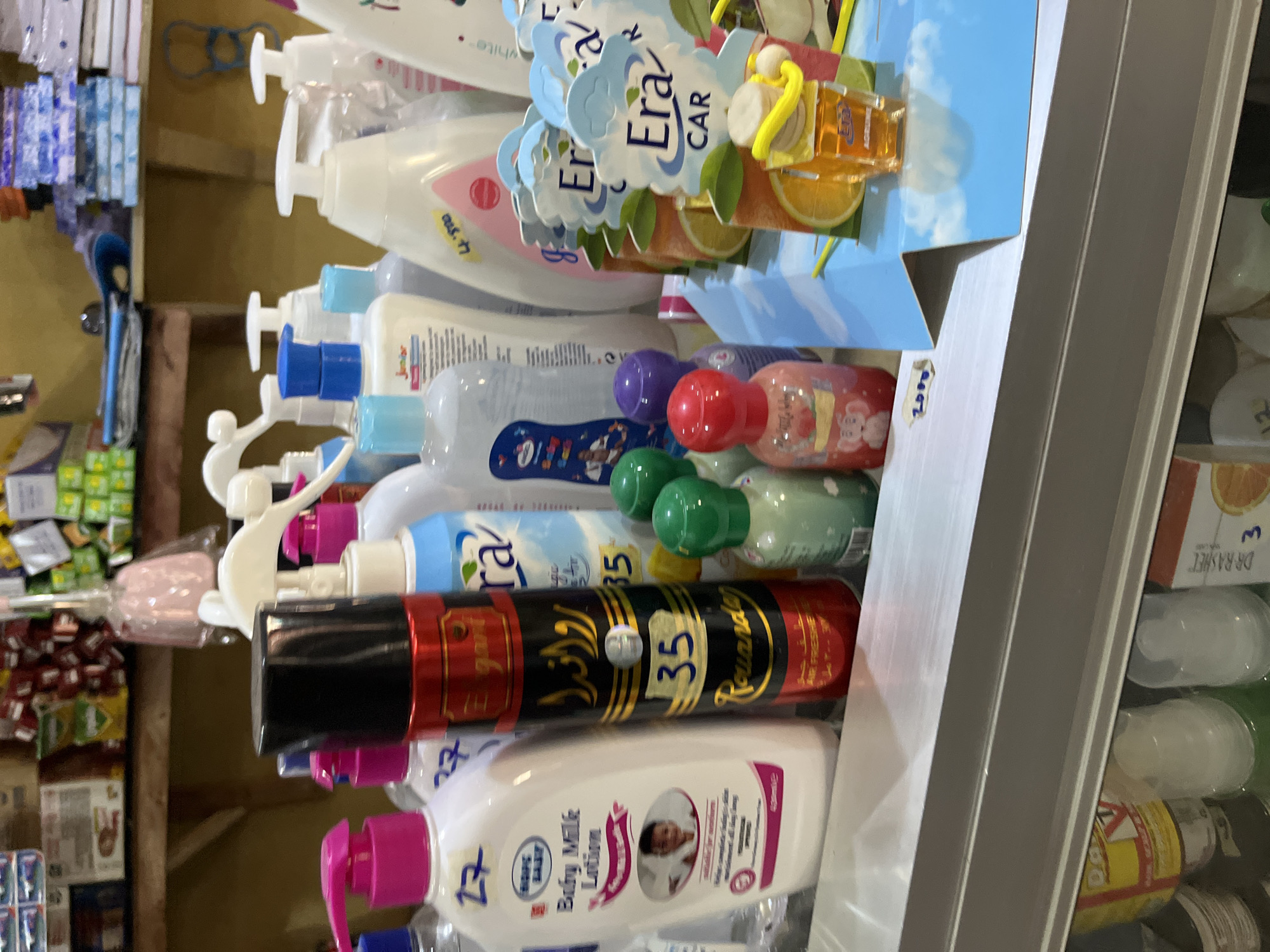 This is a forensic, matter-of-fact description of a retail display of personal care and air freshener products. The scene is likely a shelf in a small shop in Bukuru, Nigeria, given the context. The display is comprised of various plastic bottles and spray cans, predominantly featuring pink, white, and blue color schemes.
Prominently positioned in the center is a black and red spray can labeled "Rouanda" with Arabic script and the number "35" visible. To its left is a large white plastic bottle labeled "Baby Milk Lotion" with a pink pump dispenser and a sticker showing the number "27." Behind these are several other bottles, some with white pump dispensers, including a blue and white bottle with "Junior" and "27" on it, and a white bottle with a pink label featuring an image of a baby.
To the right of the central spray can are smaller, colorful bottles. These include green bottles with metallic caps, a purple-capped bottle, and a red-capped bottle with an image of an elephant. Further to the right, a display for "Era Car" air fresheners is visible, featuring graphics of oranges and lemons against a sky background. One small orange-colored air freshener is prominently displayed with a yellow hanging string.
The background shows shelves stocked with other products, including what appear to be packaged goods and possibly more personal care items. The overall environment suggests a commercial setting with a diverse range of products for sale. The lighting is moderate, illuminating the products on display. There are no individuals depicted in the image.