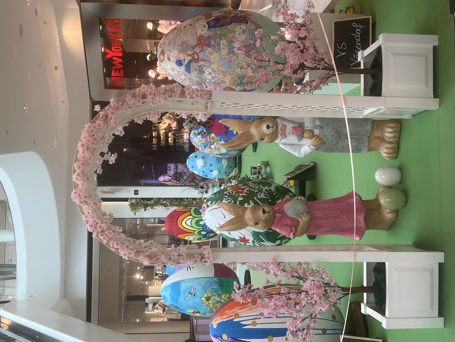 The image depicts an indoor Easter-themed decorative display located within a multi-level shopping mall in Vösendorf, Austria. The central feature is a white arched trellis, adorned with artificial pink cherry blossoms, framing a pair of anthropomorphic rabbit statues. The rabbit on the left, rendered in a brownish hue, wears a pink dress and holds a grey egg. The rabbit on the right, also brownish, is dressed in a white shirt with grey suspender pants and a pink bow tie, holding a pink rose. Two smaller, solid-colored eggs (one light green, one white) rest on the green floor covering between the statues.
The display area, covered in an artificial green surface, is populated by multiple large, egg-shaped sculptures, each uniquely painted. Examples include designs featuring Winnie the Pooh and a bee, Hello Kitty, abstract rainbow patterns, various cartoon animals, and floral motifs. Two small white planter boxes, each containing an artificial cherry blossom tree, flank the base of the arch. A light-colored ribbon or string horizontally traverses the foreground of the display. A small blackboard sign, partially visible near the right planter, bears the white chalk text "VS Vösendorf".
In the background, the architecture of a modern shopping center is evident, with expansive glass storefronts. A "NEW YORKER" store sign in bright red is prominently displayed on the right. Reflections in the glass storefronts reveal further retail interiors, mannequins, and indistinct figures, including a female figure visible through a distant storefront. Overhead lighting and curved white structural elements of the mall are also visible.