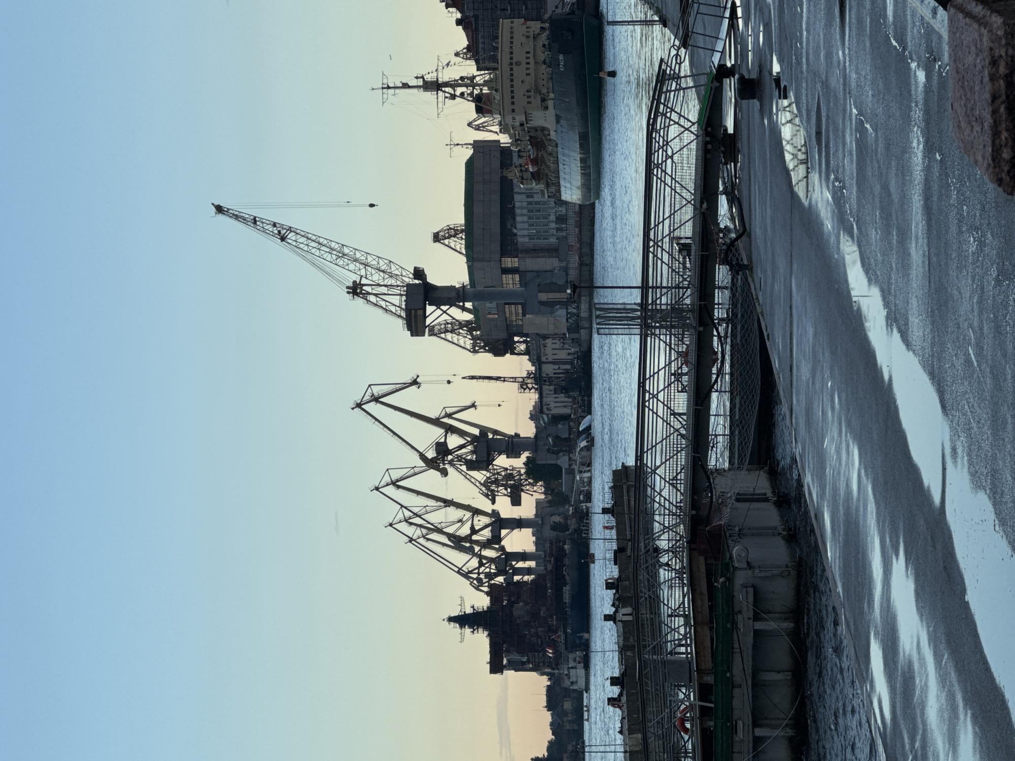 The image captures a view of the Admiralty Shipyard in Saint Petersburg, Russia, along the Neva River. In the foreground, a wet, paved waterfront area shows reflections of the sky and structures. A walkway with metal railings leads towards the water. In the middle ground, a body of water separates the waterfront from the shipyard. Several large industrial cranes of varying sizes dominate the background, silhouetted against a pale blue sky. To the right, a ship, possibly an icebreaker due to its sturdy design and markings (the name "КРАСИН" or "KRASIN" is visible), is docked. To the left of the ship, a massive, unfinished or decommissioned vessel, possibly a battleship, is under construction or renovation, with its skeletal structure exposed. The overall scene conveys a sense of industrial activity and maritime infrastructure.