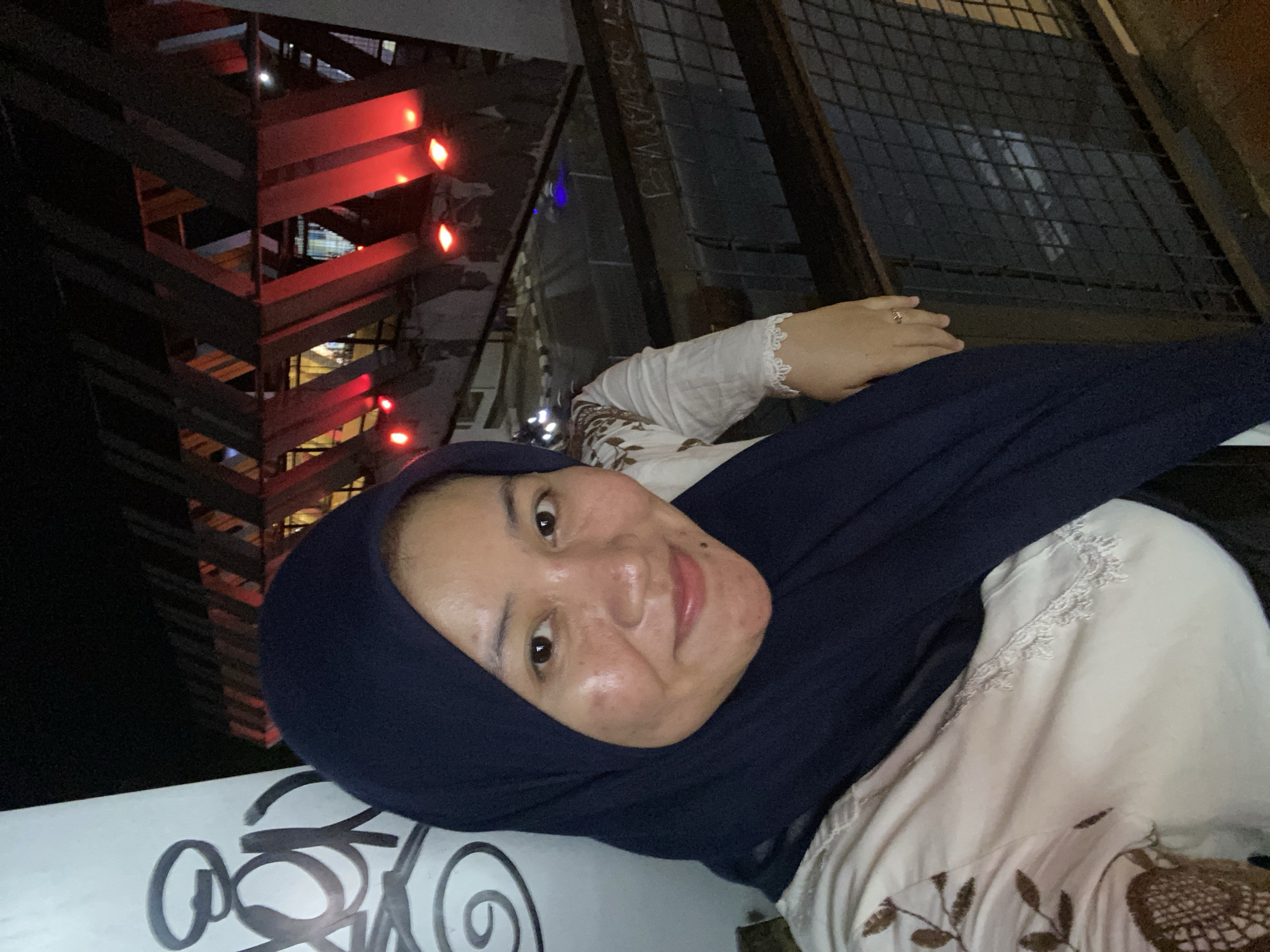 A woman, wearing a navy blue hijab and a white blouse with brown embroidery and lace trim, stands in front of a modern architectural structure at night. The structure features angled wooden beams and red lighting, suggesting it may be an outdoor event space or a bridge with decorative elements. Graffiti is visible on a white pillar to the left of the woman. The background shows a dimly lit walkway or street with some vehicle lights. The overall impression is of an urban nighttime setting in Banjarbaru, Indonesia, possibly a public space designed for aesthetics and illumination.