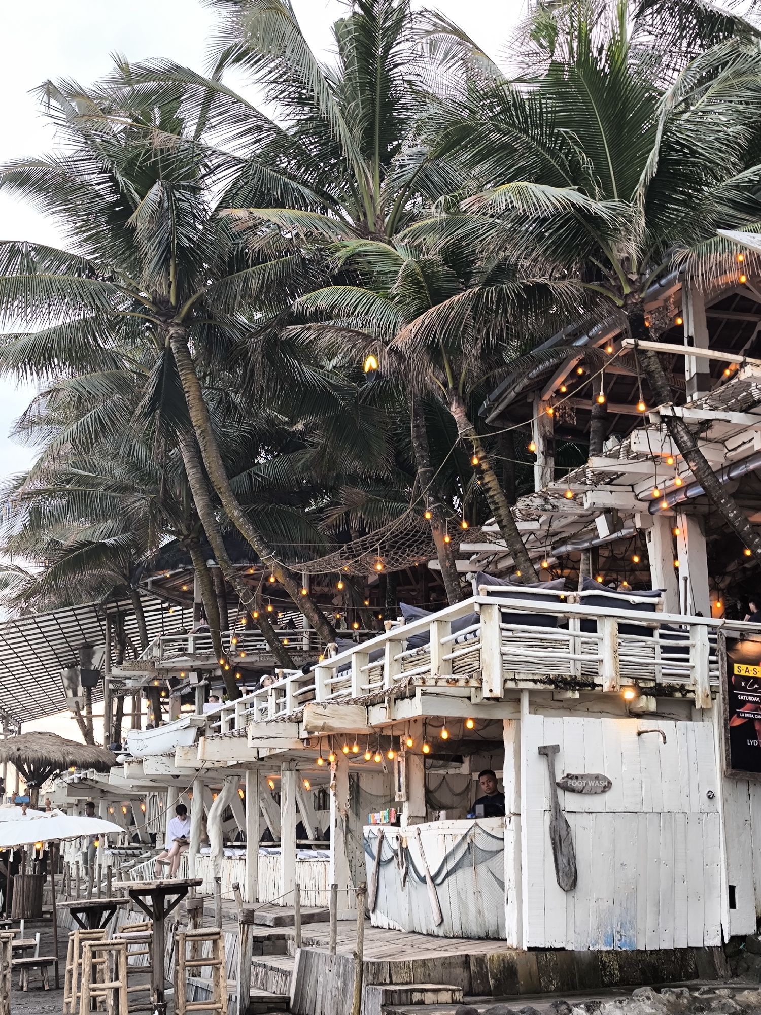 This is a medium shot, eye-level, outdoor, daytime photograph of a multi-level, open-air beach bar with a thatched-roof section on the left, in North Kuta, Indonesia. The structure is constructed primarily of white-painted wood, with bamboo accents. Several palm trees with dark green fronds are interspersed throughout the scene, with their trunks growing up through the structure. Numerous strings of warm, glowing Edison-style bulbs are strung between the palm trees and along the railings and edges of the building, illuminating the space.
The foreground shows a weathered, wooden staircase leading up to the bar's main level. To the left of the stairs are several wooden tables and chairs, some with rough-hewn finishes and others more refined. The bar itself appears to be several levels high, with white wooden railings and white lattice work on the balconies. On the lowest level, there's a bar counter visible with a man in a black shirt standing behind it, attending to customers. A sign on the side of this level reads "FOOT WASH" with an oar decoration.
On the levels above, lounge-style seating with dark cushions is visible on balconies. People are seated at these areas, appearing to be patrons of the bar. A woman in a white shirt is sitting on a ledge on one of the levels, looking down. On the far right, a portion of a black sign with yellow text is visible, listing event details like "SAS" and "SATURDAY". The ground beneath the structure appears to be a mix of sand and possibly dirt, with rocks visible at the very bottom edge of the frame, suggesting it's close to the water. The sky is overcast with a light gray hue.