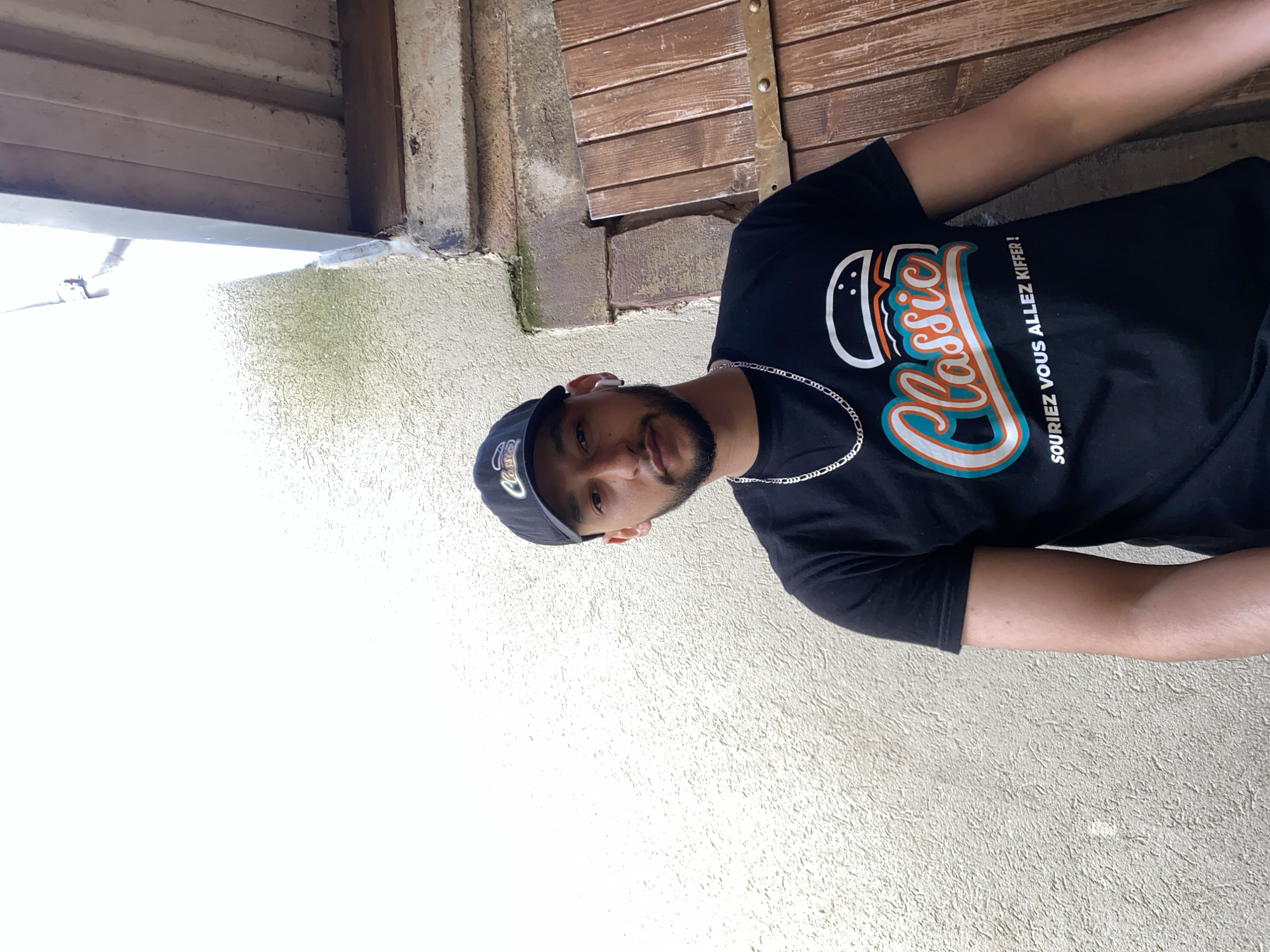 A portrait-style photograph captures a young man outdoors against a textured wall and wooden background in Schiltigheim, France. The man, with a short beard, is looking directly at the camera with a neutral to slightly smiling expression. He is wearing a black baseball cap, which also features the word "Classic" in a white stylized font. He sports a black short-sleeved t-shirt with a prominent graphic of a burger icon above the word "Classic" in a stylized orange and blue font. Below this main logo, in smaller white text, it reads "SOURIEZ VOUS ALLEZ KIFFER !". A silver chain necklace is visible around his neck, and a white earbud is in his right ear.
The background consists of rustic wooden planks, resembling shutters or a wooden wall, with some metal hardware visible. To the left, a light-colored, textured wall, possibly stucco or concrete, shows some patches of green moss or discoloration. The lighting is bright and even, suggesting it is daytime.
