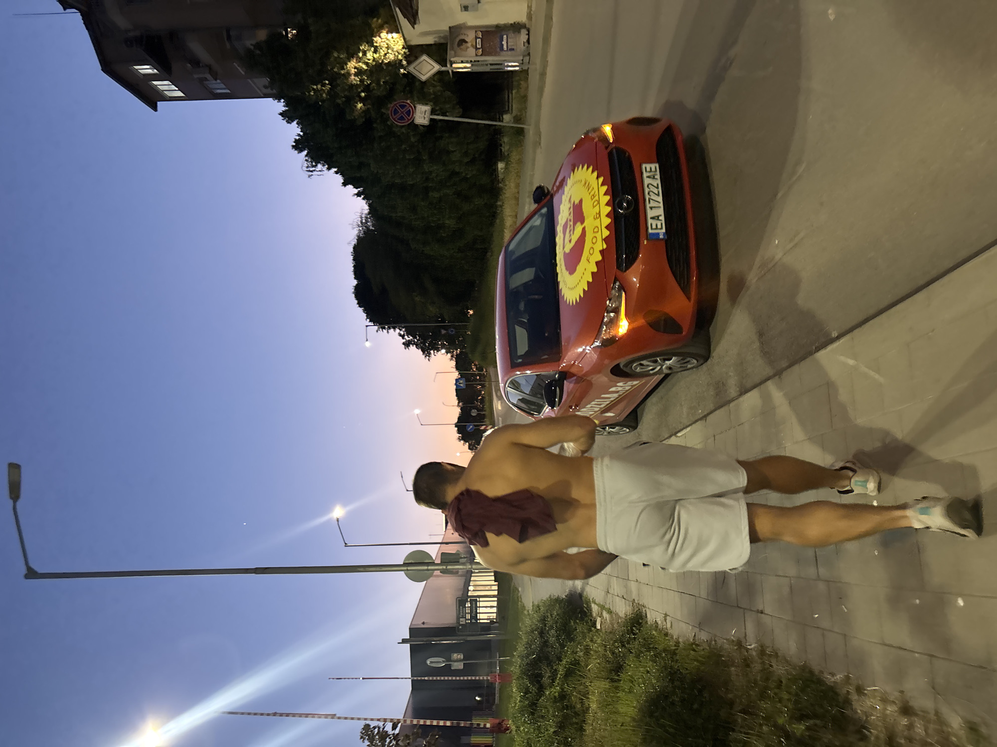 A man is walking on a sidewalk next to a street in Varna, Bulgaria, in the early evening. He is shirtless, wearing light-colored shorts and athletic shoes, with a maroon towel draped over his shoulders. Beside him, a red car with a logo on its hood is driving on the street. The car has a license plate that reads "EA 1722 AE". The sky is a deep blue with a hint of orange on the horizon. Streetlights are on, illuminating the area. Buildings and trees are visible in the background.