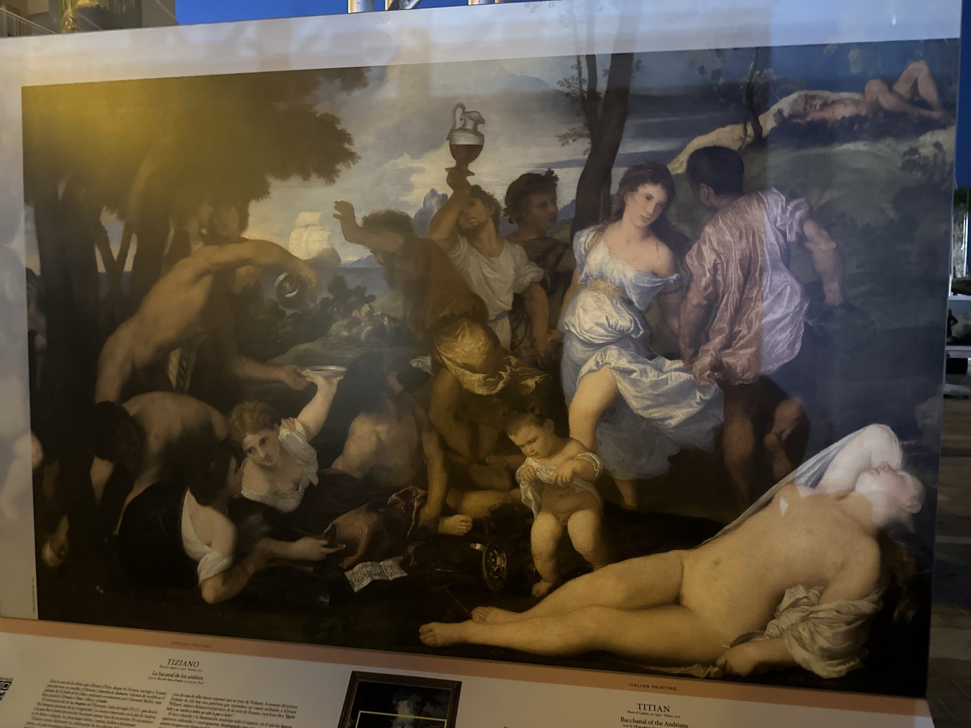 The image shows a large reproduction of Titian's painting, "Bacchanal of the Andrians," displayed outdoors in Mazarrón, Spain. The painting depicts a chaotic scene of revelry with numerous figures, both male and female, engaged in drinking, dancing, and embracing. Nudity is prevalent throughout the artwork, with many figures being scantily clad or completely undressed. The central figures include a reclining nude woman, a group of men holding a jug and cup, and a child. The background features a pastoral landscape with trees and a body of water. The reproduction is mounted on a flat surface and appears to be part of an exhibition, as suggested by the text at the bottom of the image which identifies the artwork and artist. The lighting in the image indicates it was taken at dusk or during the evening, with artificial light illuminating the reproduction.
