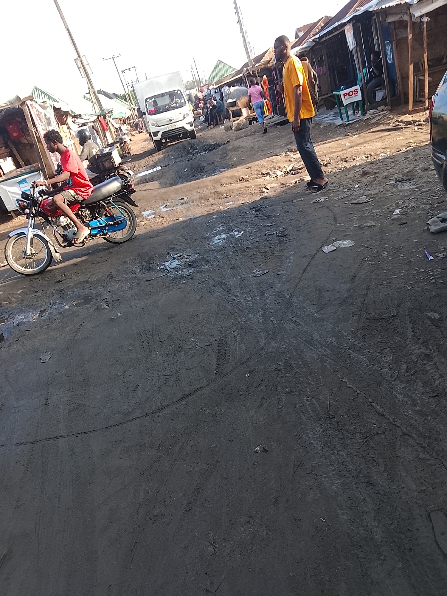 The image depicts an unpaved street in what appears to be a market area in Abuja, Nigeria. The street is muddy and shows tire tracks. A man wearing an orange shirt and dark pants stands on the right side of the frame. To his left, a man on a blue and red motorcycle is riding, barefoot. In the background, several people walk along the street and near makeshift shops. A white van is visible. A "POS" sign is attached to one of the shops.