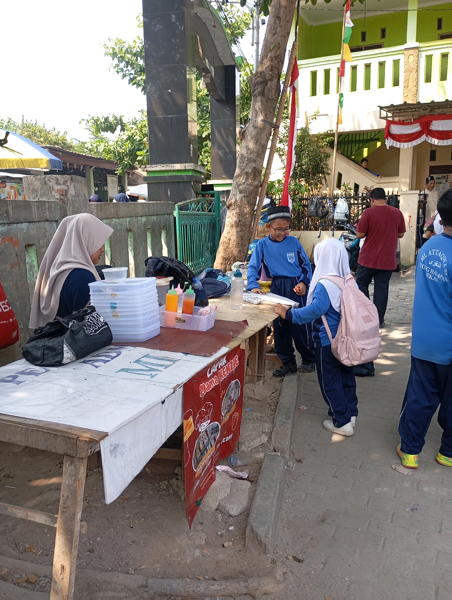 In Babelan, Indonesia, a street vendor wearing a hijab is serving customers from a roadside stall. The stall has a white tablecloth spread over a wooden table, laden with food containers and condiment bottles. Behind the vendor, a stack of white plastic containers is visible. The street is paved with bricks, and a small concrete barrier runs along the edge. In the background, a green building with white window frames stands tall, decorated with red and white bunting. Children in blue uniforms, presumably students, are seen interacting with the vendor and each other. One child, wearing a white hijab, carries a pink backpack. The atmosphere appears to be that of a bustling marketplace on a sunny day.