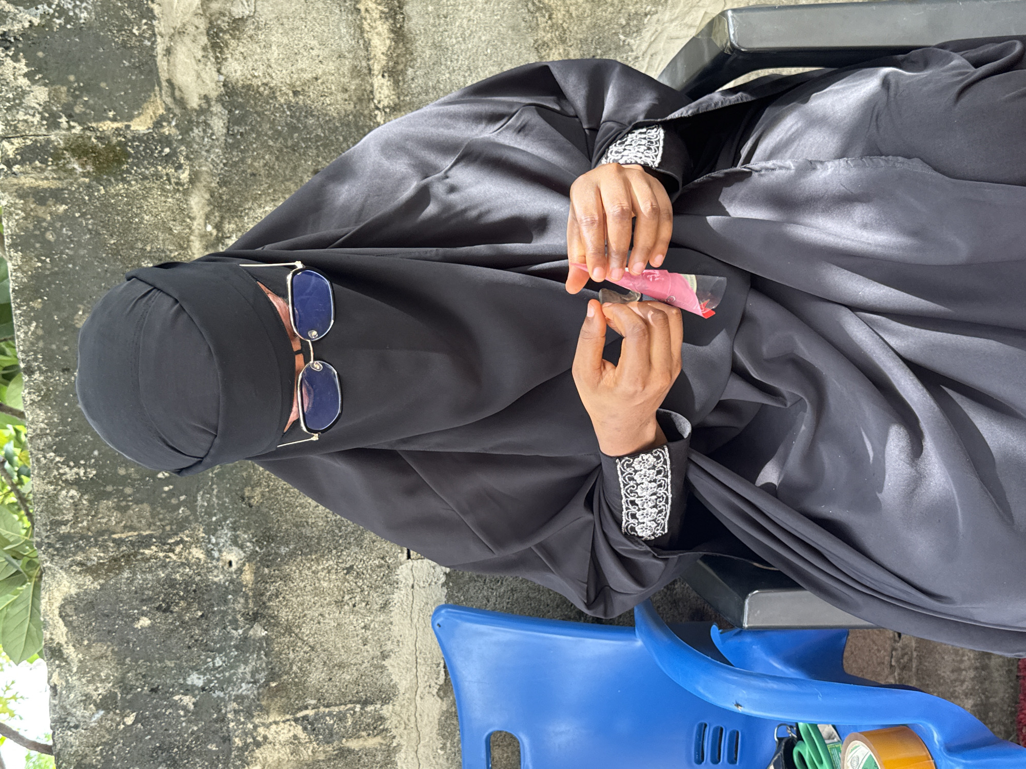 The image captures a person, likely female, seated outdoors in Ado, Nigeria, under bright daylight conditions. She is wearing a black niqab, covering her face except for her eyes, and a flowing black abaya. Blue-lensed sunglasses with gold frames are resting on her niqab. Her hands, which are visible and have a dark skin tone, are holding a small pinkish or reddish package, possibly a sachet or tube. The sleeves of her abaya feature prominent white and black embroidered cuffs with a floral or lace pattern.
The person is seated on a black plastic chair, which is partially visible. In the foreground, a blue plastic chair is also visible, holding various items including a pair of green-handled scissors, keys, and a roll of brown packing tape (text on tape is unreadable). The background features a textured, light grey concrete or stone wall, with hints of green foliage from a plant on the left side, suggesting an outdoor garden or courtyard setting. The ground appears to be rough concrete or paved. The bright light and clear shadows indicate a sunny daytime environment. The activity appears to be a moment of rest or preparing to use the item in her hands. No emotions are discernible due to the face covering.