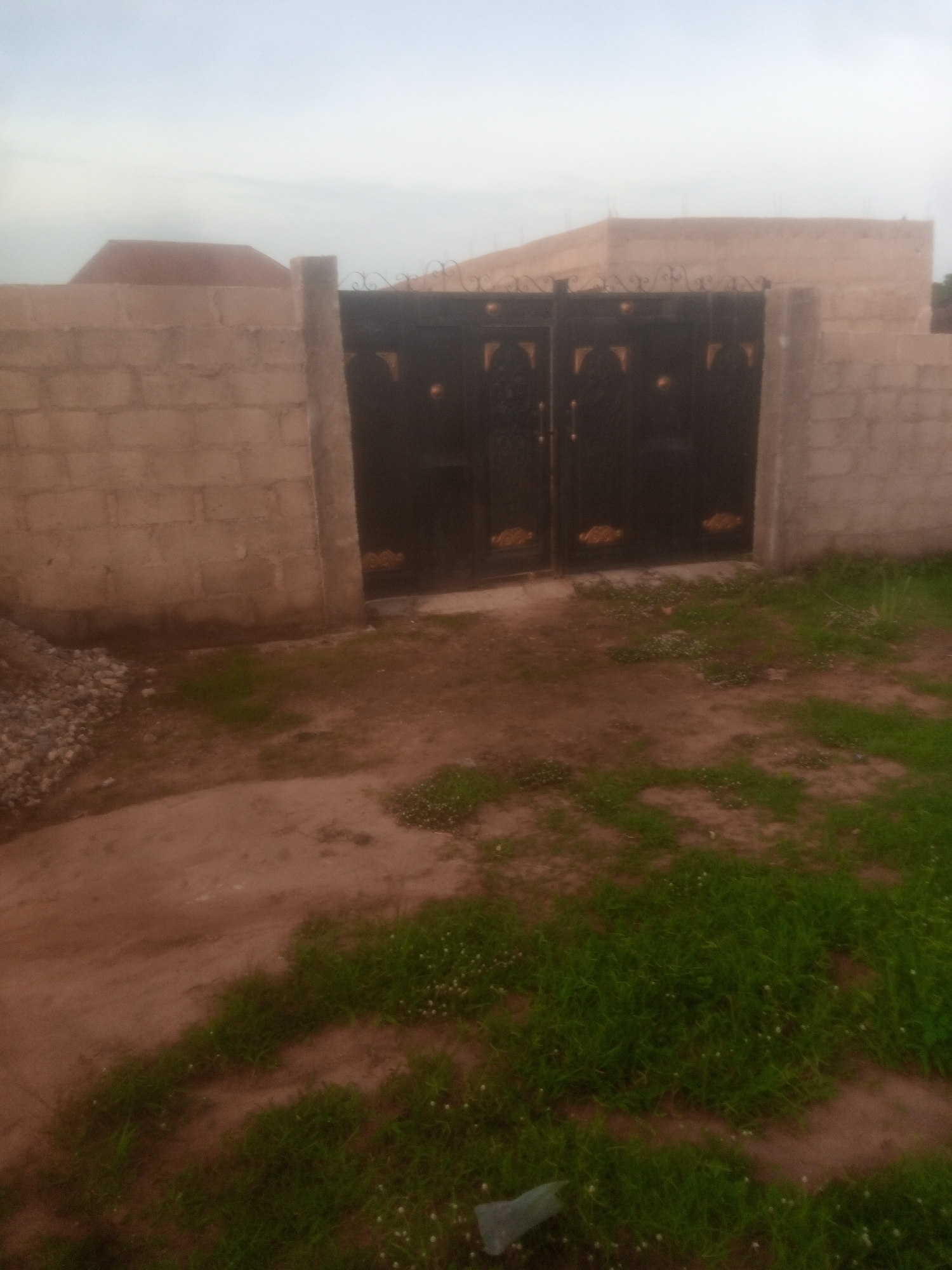The image shows a dark metal gate with gold accents, flanked by a concrete block wall. The gate is partially open, revealing a building behind it. The ground in front of the gate is covered with dirt and patches of green grass. The sky above is overcast. The scene is located in Jalingo, Nigeria.