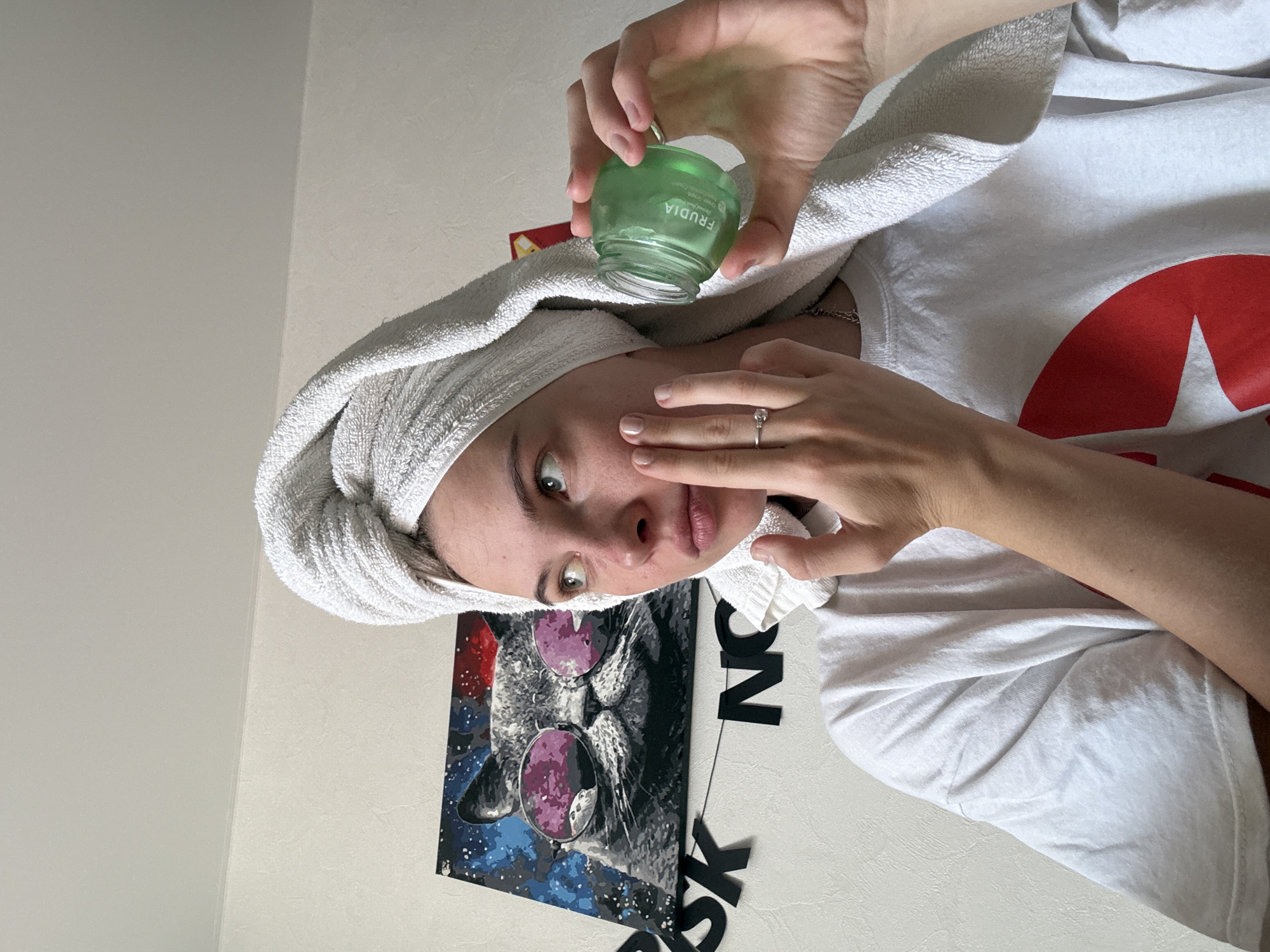 A young woman with a white towel wrapped around her head and shoulders, wearing a white t-shirt, is applying a green cream from a jar to her face. She is looking to her right, with her left hand touching her cheek and her right hand holding the opened jar. The background shows a wall with a painting of a cat wearing sunglasses, and black letter decorations on the wall. This scene is located indoors, likely in a residential setting. The specific location in the city of Perm, Russia cannot be determined from the provided image.