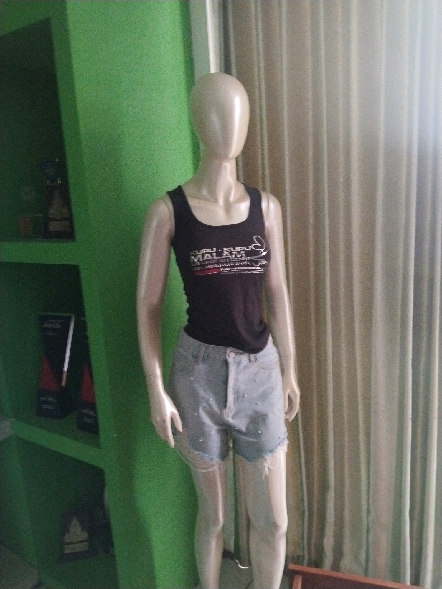 A mannequin, displaying a black tank top and light blue denim shorts, stands against a green wall and beige curtains in Sinduadi, Indonesia. The tank top has a graphic print with silver lettering that reads "MAH XUPU AM AUTO CUSTOM! 100% PENGHA RAUKIN MOBIL". The denim shorts are distressed with frayed hems and decorated with small, light-colored studs. The mannequin's pose is neutral, with its arms at its sides. To the left of the mannequin, a green shelf unit is visible, containing several framed items and other objects. The overall environment appears to be an indoor retail or display space.