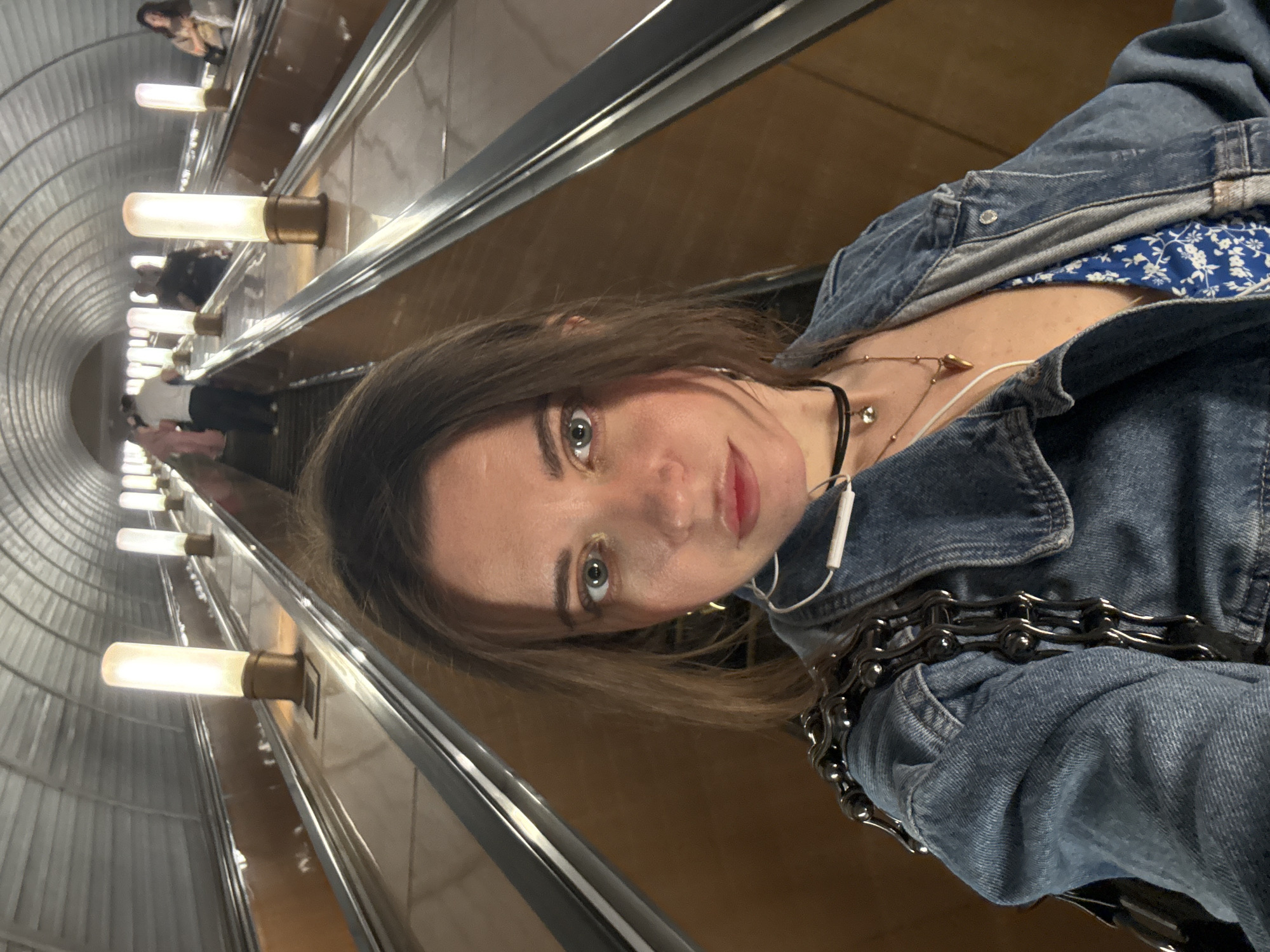 A medium shot captures a woman wearing a denim jacket and multiple necklaces, including a black corded one and a delicate gold chain. She is also wearing an earbud. A glimpse of a blue and white patterned garment is visible beneath the jacket. The woman is on an escalator in the Moscow Metro, recognizable by the distinctive tubular lighting fixtures along the escalator walls. Several people are visible in the background.