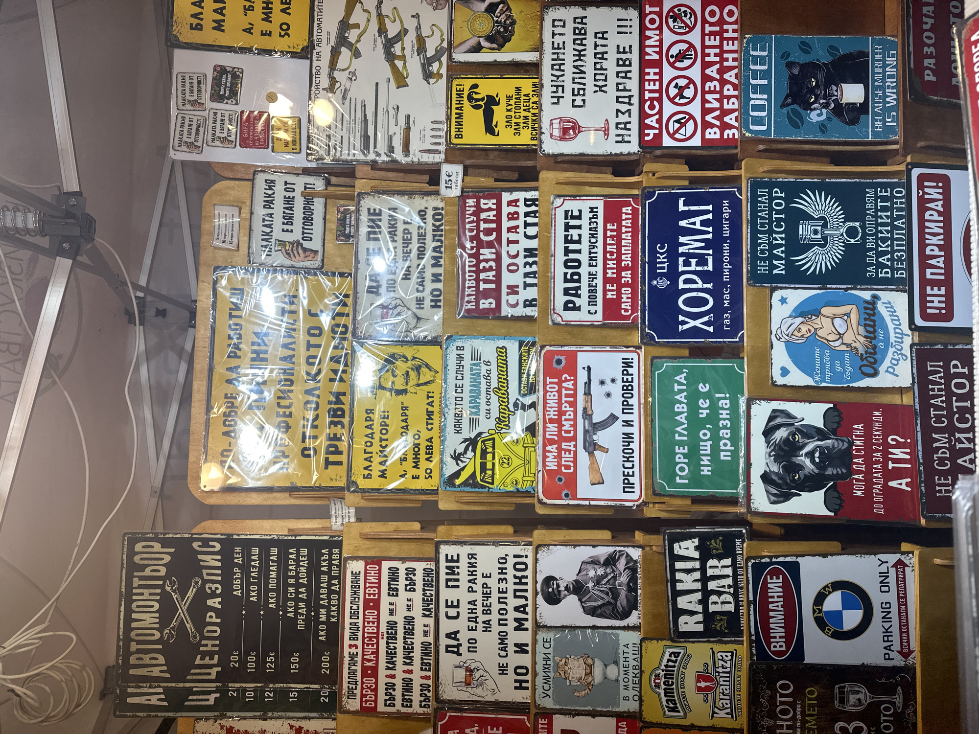 The image depicts a display of novelty metal signs for sale. The signs feature various humorous and satirical messages in Bulgarian. The signs are organized in rows, appearing to be stacked on a table or display stand. Subjects include drinking, cars, mechanics, coffee, and other themes. Textures appear aged and distressed, suggesting a vintage aesthetic. The image includes a price list in the corner, indicating prices in both Euro (€) and Bulgarian Lev. (лв).