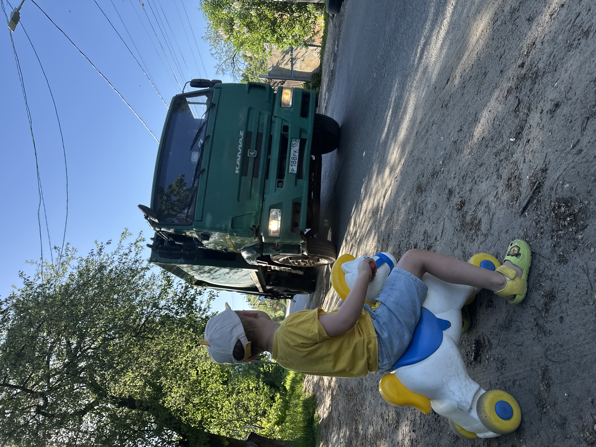 A young boy sits on a white and blue plastic toy horse with yellow wheels, facing a green Kamaz truck with the license plate P 188 PK 152, parked on a paved surface. The boy wears a yellow shirt, denim shorts, a baseball cap, and yellow sandals. The truck has its headlights on. The scene is outdoors with trees and greenery visible in the background. The location is Nizhny Novgorod, Russia.