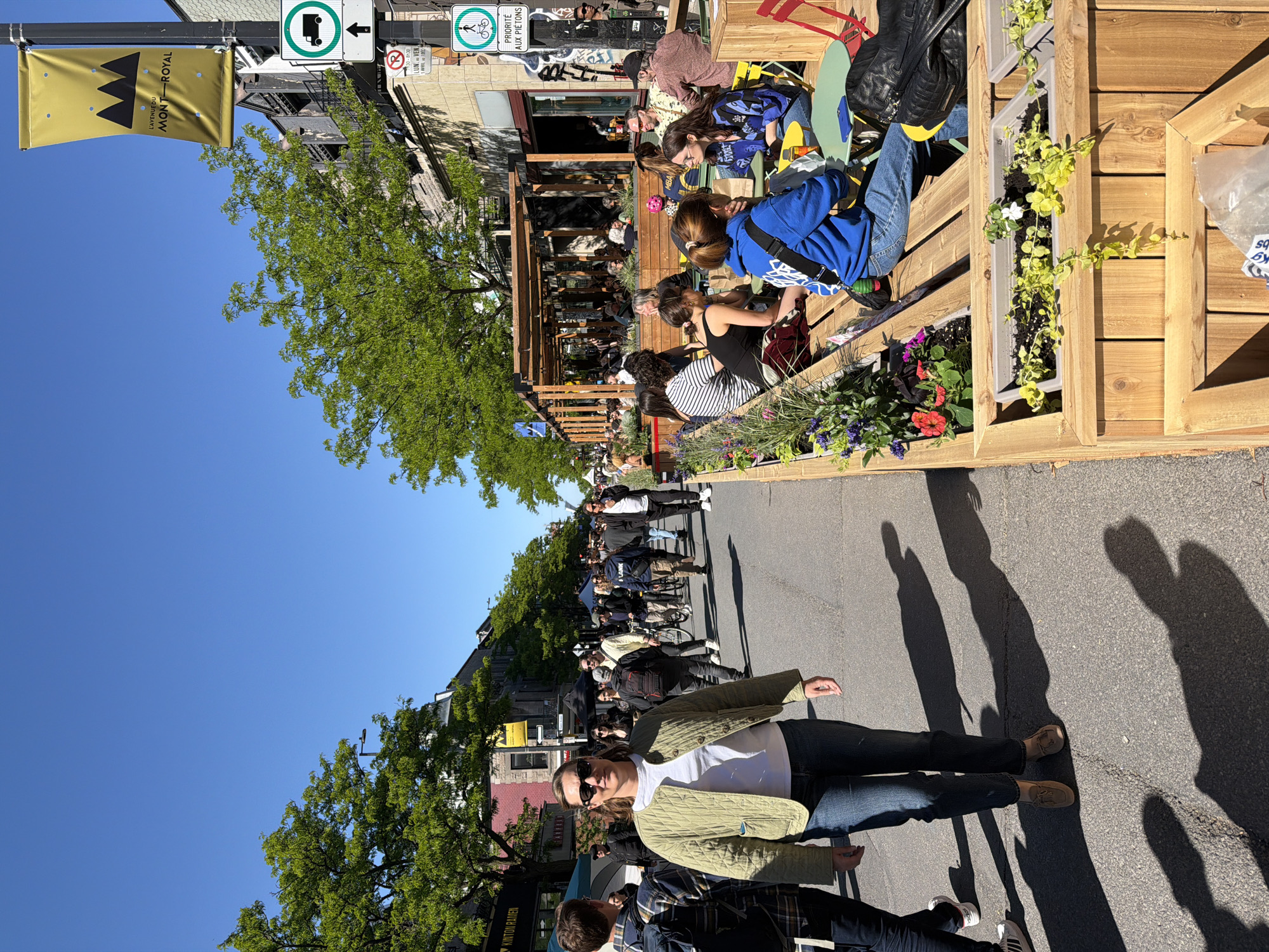 The image captures a vibrant scene on L'Avenue du Mont-Royal in Montreal, Canada. The view is along a street where an outdoor dining area has been created, constructed with wooden frames and planters filled with flowers and greenery. People are seated at tables, engaging in conversation and enjoying meals. The street is also bustling with pedestrians. A woman wearing a light green jacket and sunglasses walks along the street. Shadows of pedestrians and objects are cast on the asphalt road, suggesting bright sunlight. Street signs, including one indicating "Priorité aux Piétons" (Priority to Pedestrians), are visible. A banner bearing the name of the avenue, "L'Avenue du Mont-Royal," and the name of a restaurant "KINTON RAMEN" hangs on the pole. The overall atmosphere is lively, capturing the essence of outdoor urban life.
