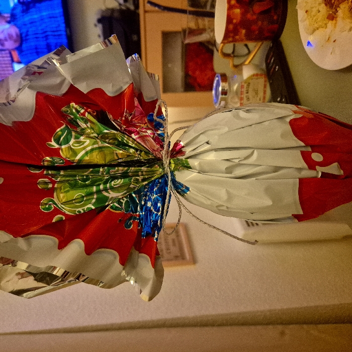 The image displays an indoor domestic scene. A large, vertically oriented object, likely an Easter egg, wrapped in decorative foil, occupies the foreground. The upper section of the wrapping is predominantly red with green, blue, and multi-colored intricate patterns, gathered and tied around the middle with a thin, silver-colored string. The lower section of the wrapping is silver foil, terminating in a red base with white shapes.
In the background, to the left, a light-colored wall is visible, featuring a partially obscured framed item with faint text including "CHROPO" and "2016." Below this, a white, rectangular object, possibly a heating unit or radiator, is partially visible. A light-colored curtain or drape runs along the far left edge of the frame.
To the right of the main subject, a wooden surface, likely a table or countertop, supports various items. In the bottom right corner, a white plate containing remnants of food, specifically a yellowish granular substance mixed with a reddish sauce, is present. Behind the plate, a dark-colored remote control lies on the surface. Further back, a small glass jar with a metal lid and a partially legible label, containing what appears to be salt, is visible. Adjacent to the jar is a red mug with a golden handle and an indistinct pattern. Beyond the table, a wooden cabinet is visible, topped by a television screen displaying a blurred image of a person, identifiable as a man in a blue shirt.
The scene depicts a residential interior. No specific visual cues are present to identify the exact location within Strasbourg, France; the environment is consistent with a generic domestic setting.
