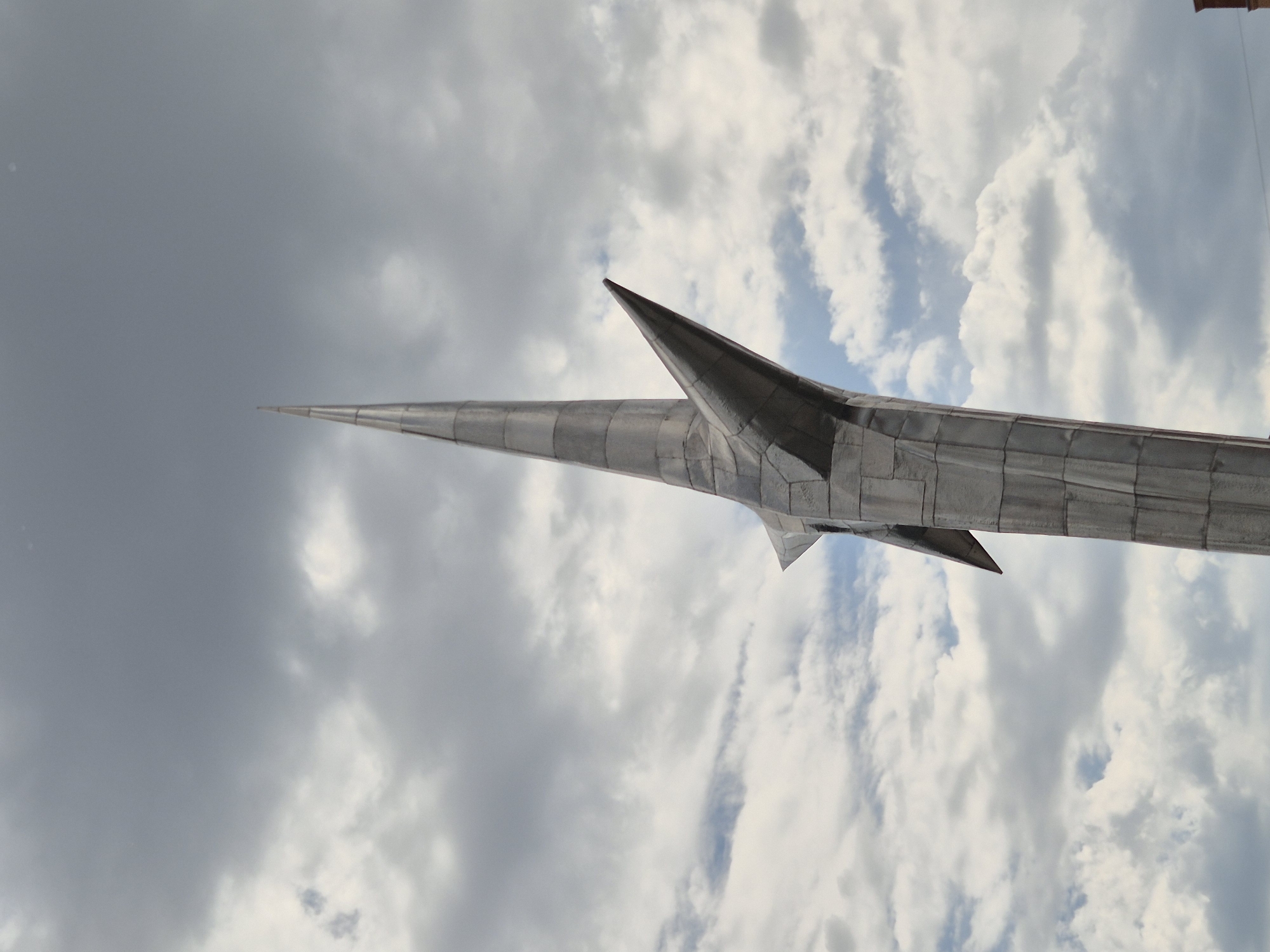 The image shows a close-up, low-angle shot of a tall, metallic spire, possibly a monument or sculpture, against a cloudy sky. The spire is segmented and appears to be made of polished metal. It has a sharp, pointed top and several angular protrusions extending outwards from its sides, resembling wings or blades. The sky is filled with a mix of dark gray storm clouds and lighter, brighter patches, suggesting dynamic weather conditions. The metallic surface of the spire reflects the light from the sky.
This structure is located in the city of Kokshetau, Kazakhstan, and is known as the Monument to the Founders of Kokshetau. It is a prominent landmark in the city.