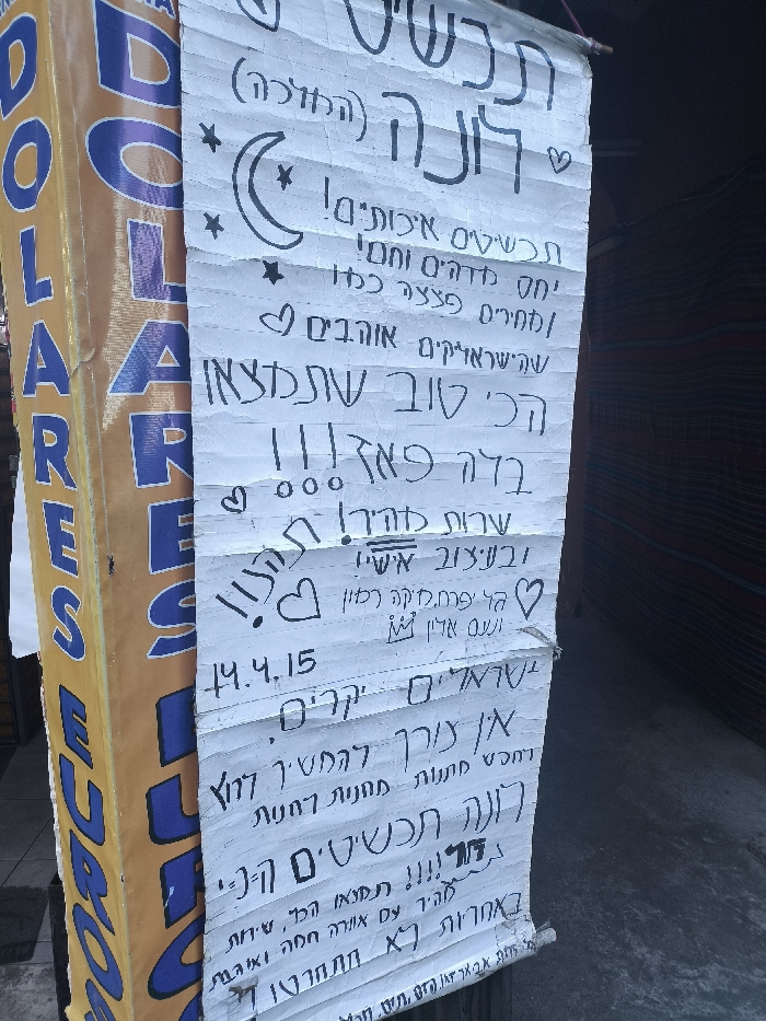A hand-written sign in Hebrew advertises "Luna Jewelry". It claims the jewelry is of high quality, offers a wonderful and warm attitude, and exchanges at a great rate for Israelis. It suggests it's the best place to find jewelry, with fast service and special designs. The sign also has the date 14.4.15, a drawing of a crescent moon and stars, and several hearts. It also states that you won't regret coming, as the service is warm and loving. The sign is hung next to a store called "DOLARES EUROS".