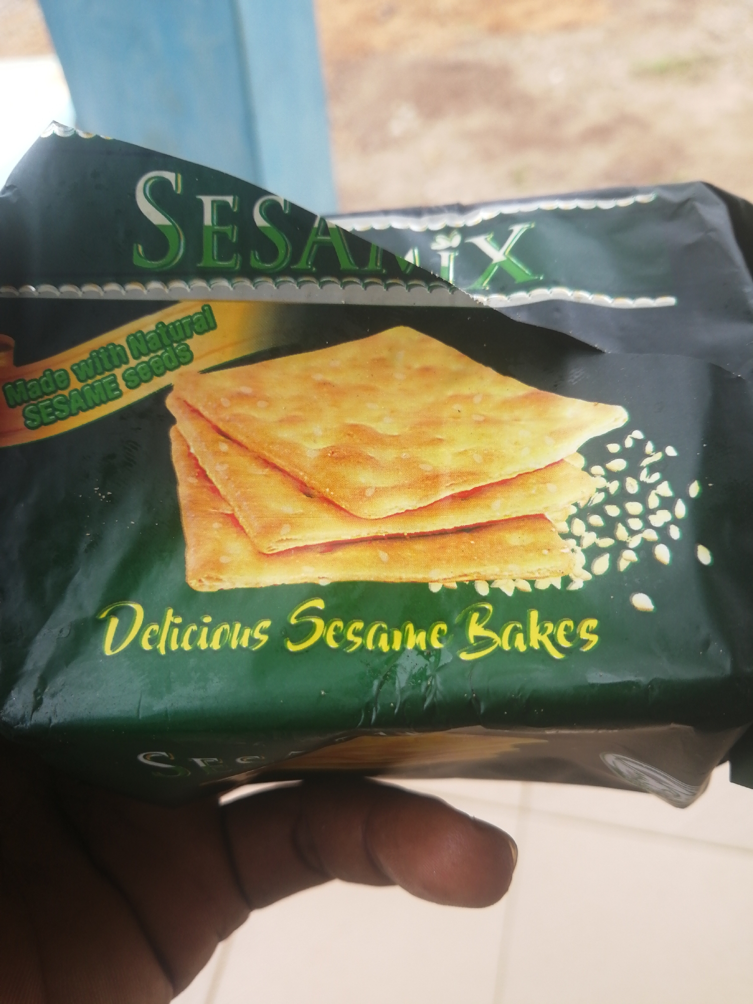 An open package of SESA-MIX crackers is held in a person's left hand in Ajasse Ipo, Nigeria. The package is primarily green with a light blue and yellow logo. The crackers are golden brown and stacked on top of each other, with a scattering of sesame seeds around them. The text on the package reads "SESA-MIX" at the top, "Made with Natural SESAME seeds" in the middle, and "Delicious Sesame Bakes" at the bottom. The background is blurred and out of focus, but appears to be an outdoor setting with a light blue structure and a brown, dry ground. The person's hand is dark-skinned and only their fingers and thumb are visible, holding the bottom edge of the package.