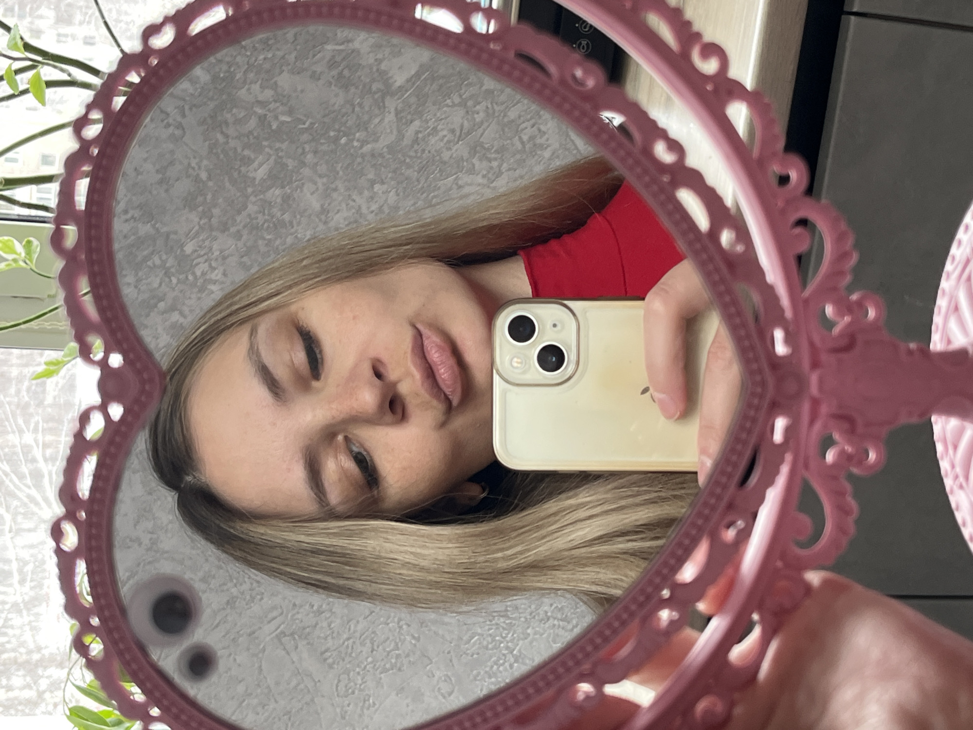 A woman is taking a selfie in a heart-shaped pink mirror. She is wearing a red shirt and holding a light gold iPhone. The background visible in the mirror includes a gray textured wall and a window with a plant outside. The location is likely Chaikovsky, Russia, although there are no specific landmarks or indicators of this location.