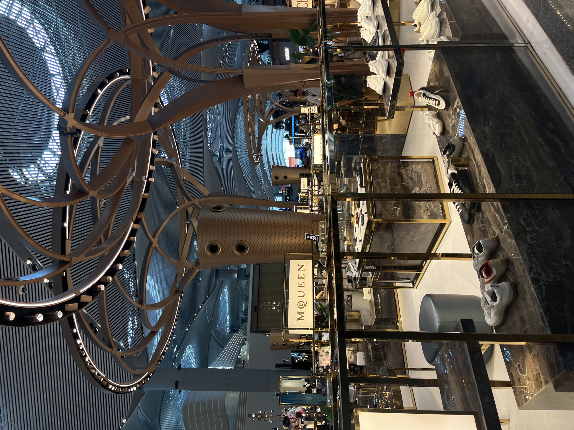 The image depicts the interior of a modern retail establishment, likely a high-end shoe store, within a large, architecturally significant building. The store features polished marble display cases with gold accents, showcasing various footwear, predominantly sneakers. In the background, a sign reads "MQEEN". The ceiling of the space is a prominent feature, with a complex, sculptural design of interwoven wooden beams and linear lighting. The overall ambiance is one of luxury and contemporary design. Given the context provided, this scene is located within the Istanbul Airport in Arnavutköy, Turkey. There is one male individual visible in the background, appearing to be a store employee or security personnel, standing near a display of footwear. The environment is well-lit, with a mix of natural light filtering through the ceiling structure and artificial lighting.