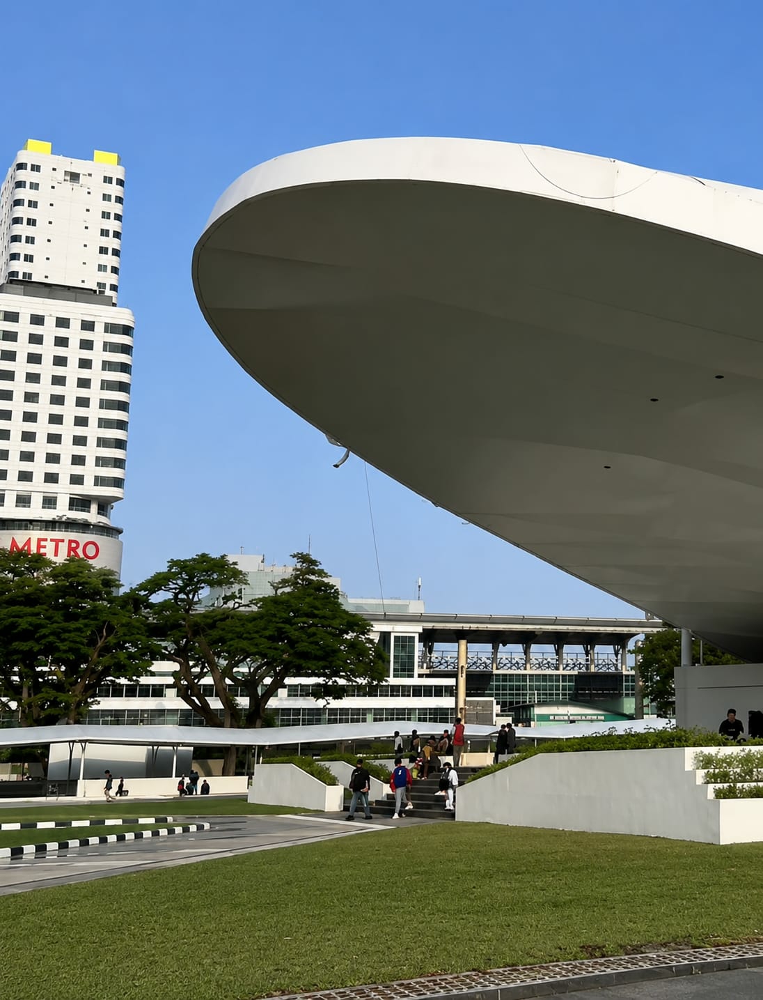 The image displays a modern architectural scene in the City of Medan, Indonesia. In the foreground, a large, curved white overhang dominates the right side of the frame. Below it, a lush green lawn is visible, with a pathway marked by black and white striped borders in the lower left corner. A set of white tiered stairs leads up to the overhang, and several people are ascending or descending them.
In the background, a tall, white rectangular building with numerous dark windows and yellow accents at the top stands prominently. The word "METRO" in red letters is visible on the side of this building. To the left of this tall building, a cluster of large, green trees are situated in front of a white, multi-story building with a linear architectural design and dark-framed windows. Further in the background, another building with a skywalk or elevated walkway can be seen. The sky is clear and blue. The overall impression is one of a contemporary urban environment, possibly a public space or a corporate complex.