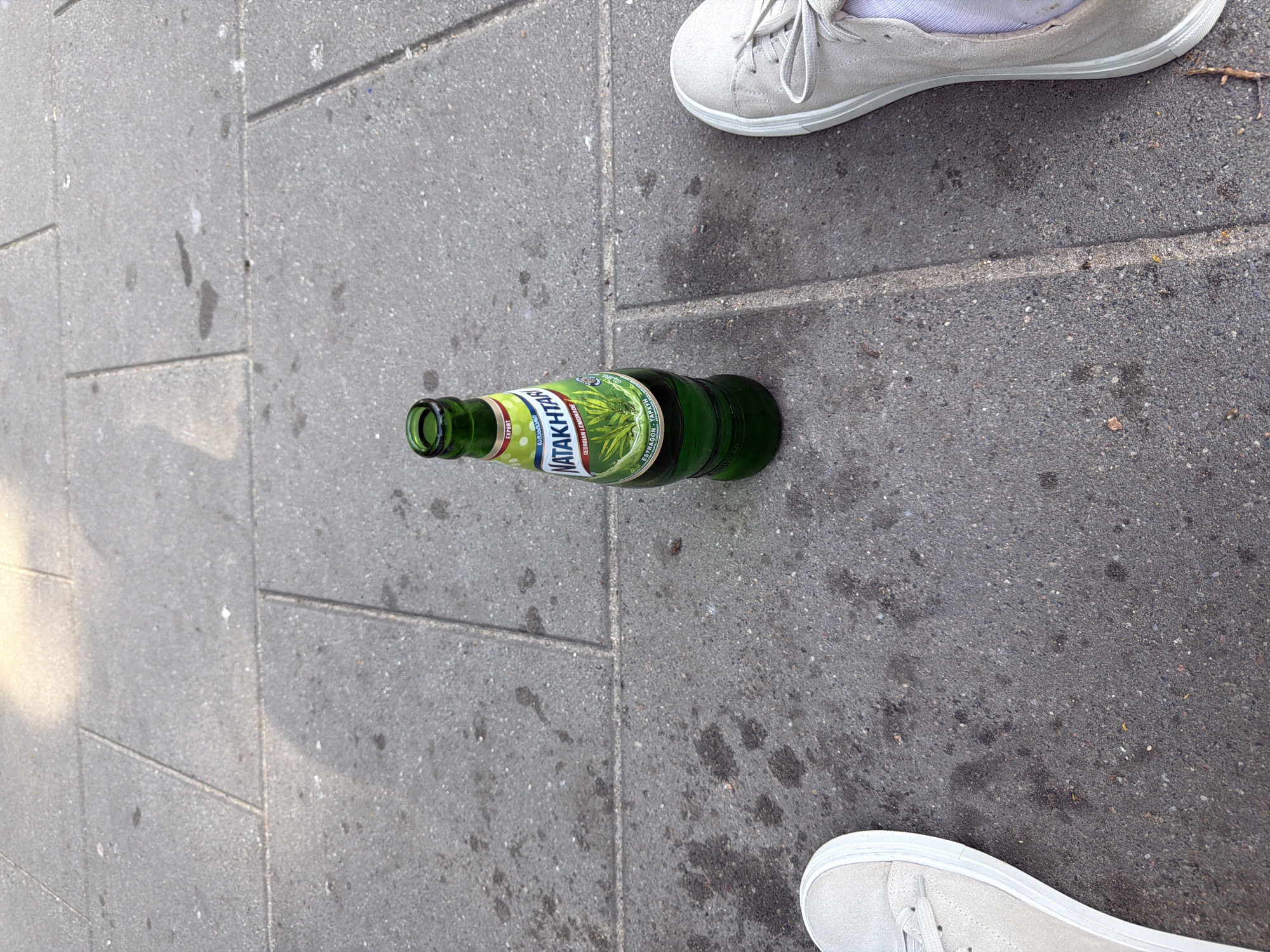 The image shows a close-up, eye-level view of a green glass bottle of Natakhtari Georgian Lemonade with an "ESTRAGON - TARKhUN" label, standing upright on a gray tiled sidewalk. Two pairs of feet wearing light-colored canvas shoes are visible, one on the left and one on the right, suggesting two people are sitting or standing near the bottle. The sidewalk is composed of rectangular tiles with visible grout lines. Sunlight casts shadows on the ground.