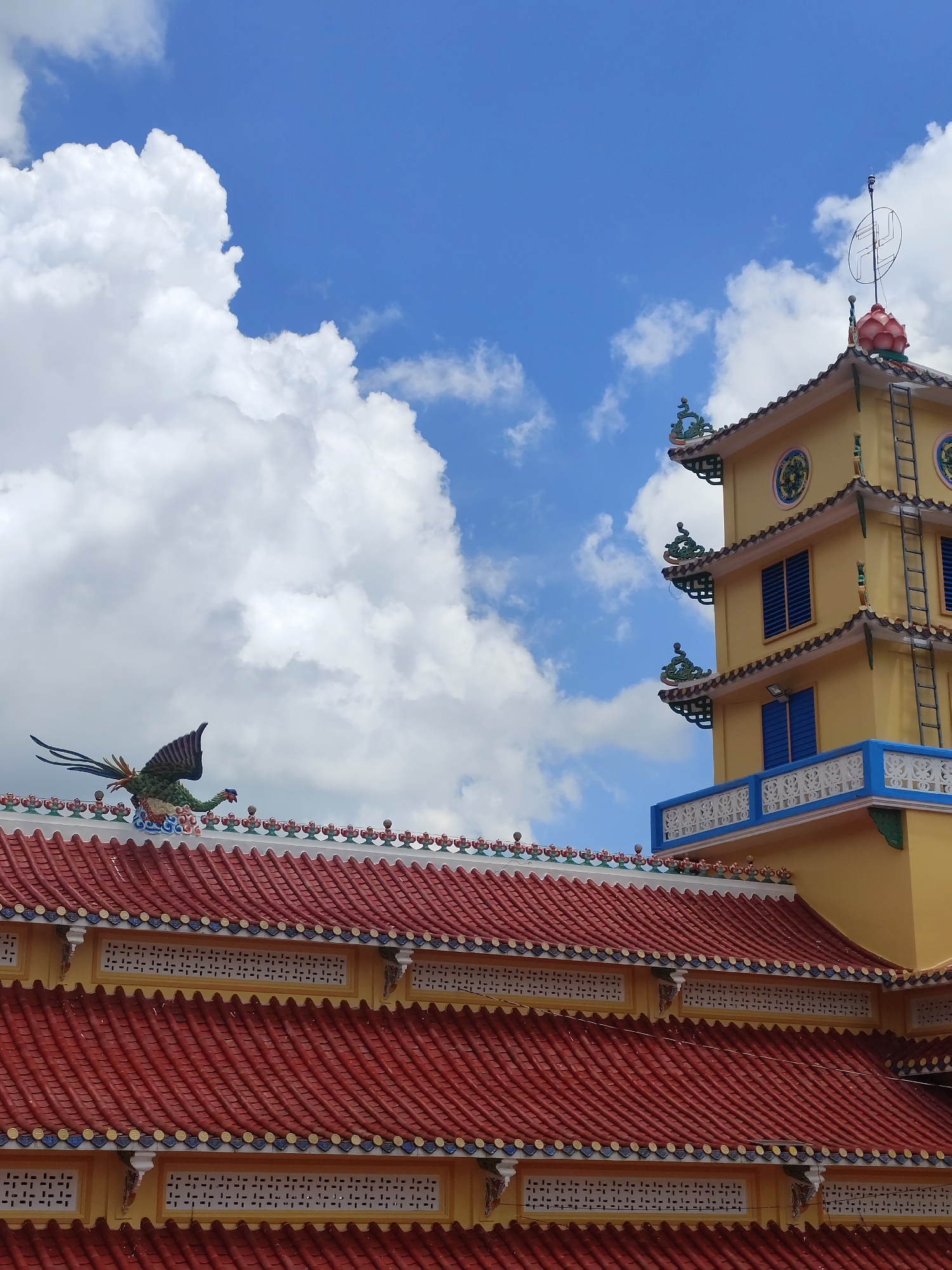 The image shows a section of a yellow temple with a red tiled roof in Lộc Quý, Vietnam. The temple features multiple tiers, decorative eaves, and blue-shuttered windows. A ladder is visible on the side of the tower. A green, dragon-like statue stands on the ridgeline of the lower roof. The sky is blue with scattered white clouds. The foreground is dominated by the red tiled roof with decorative elements.
